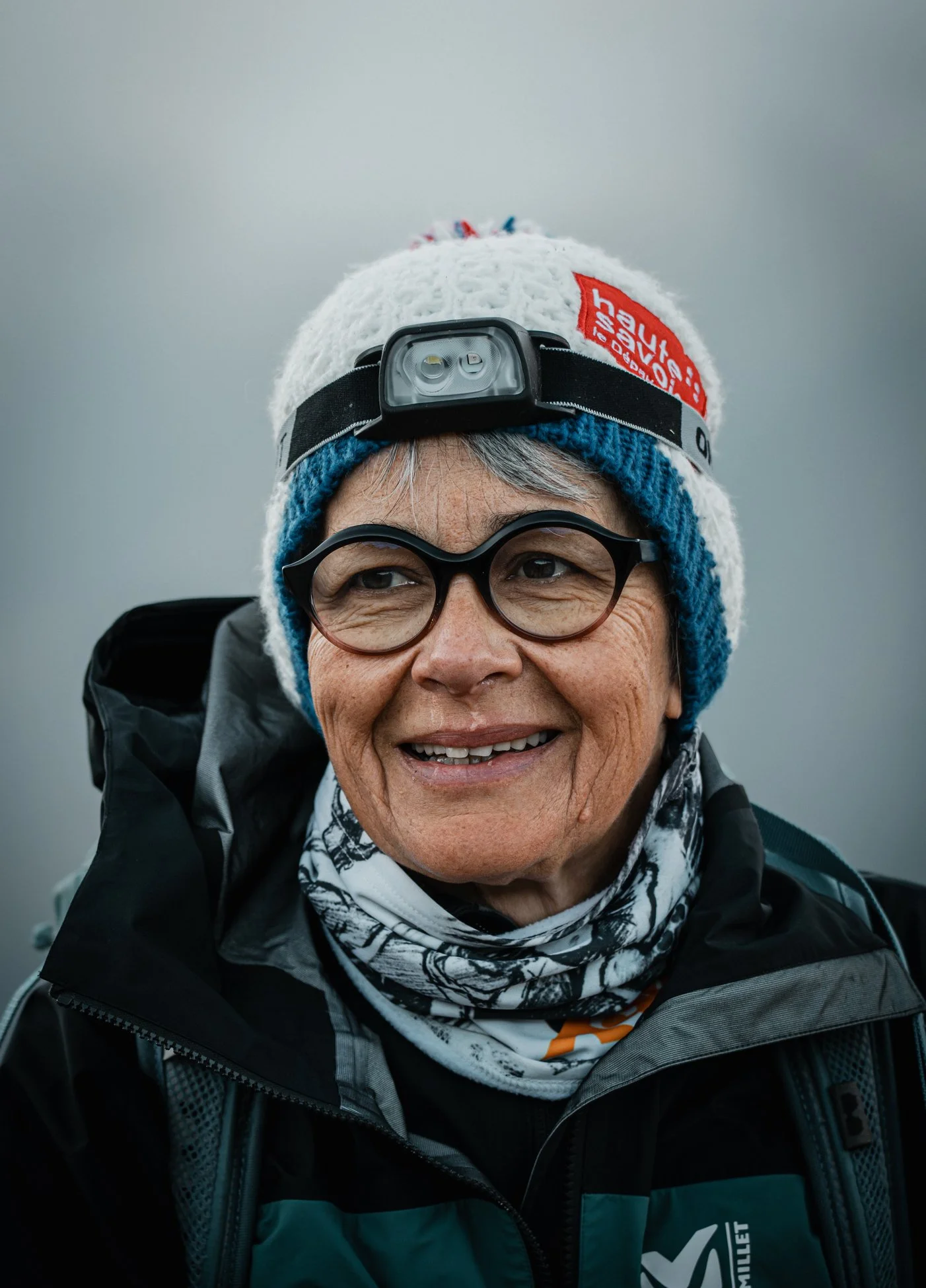 Une femme âgée portant des vêtements de randonnée, une écharpe, une casquette avec une lampe frontale et des lunettes, souriant dans un environnement brumeux.