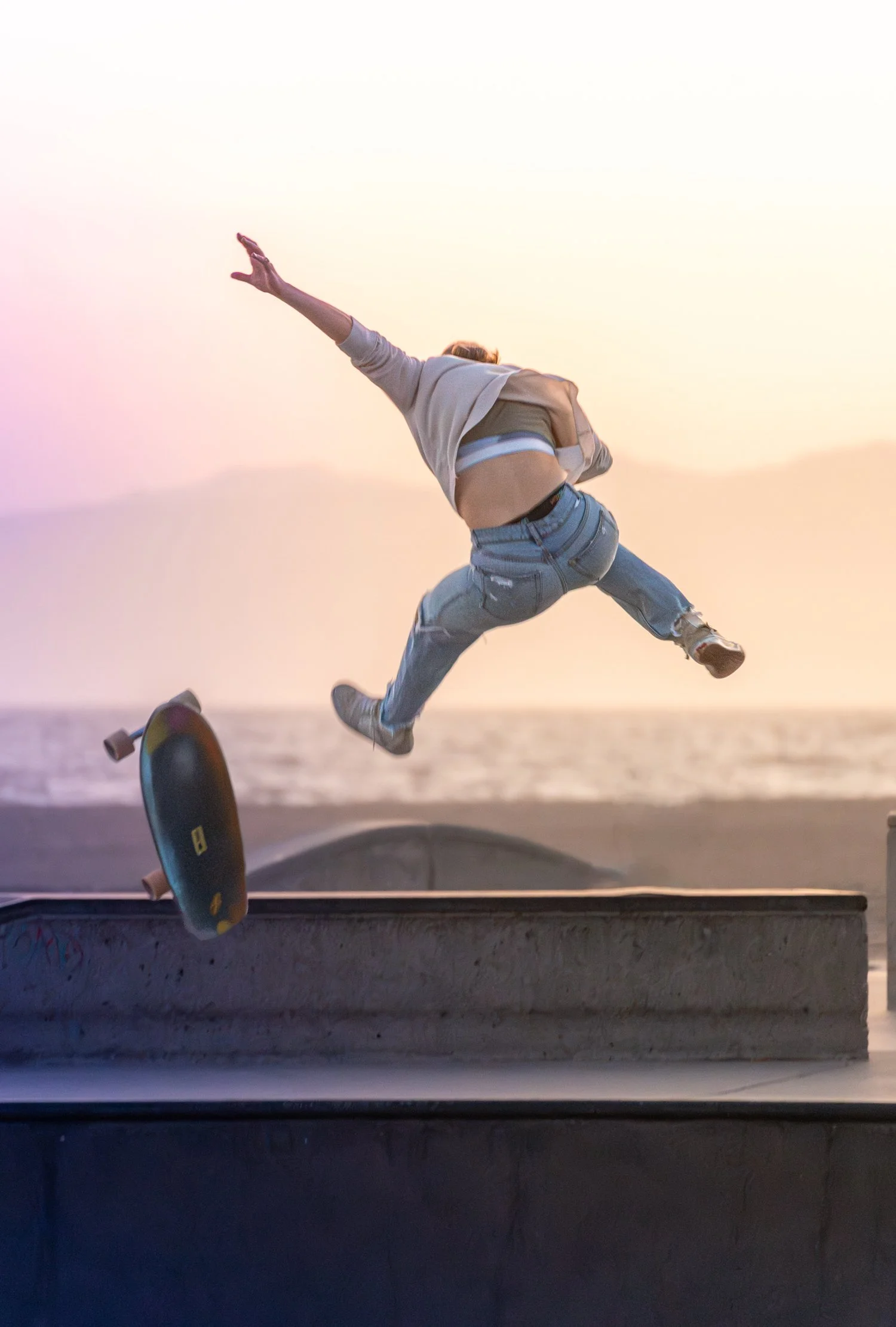 Jeune femme en jean déchiré effectuant une saut en skateboard au crépuscule, vue de derrière, à la plage.