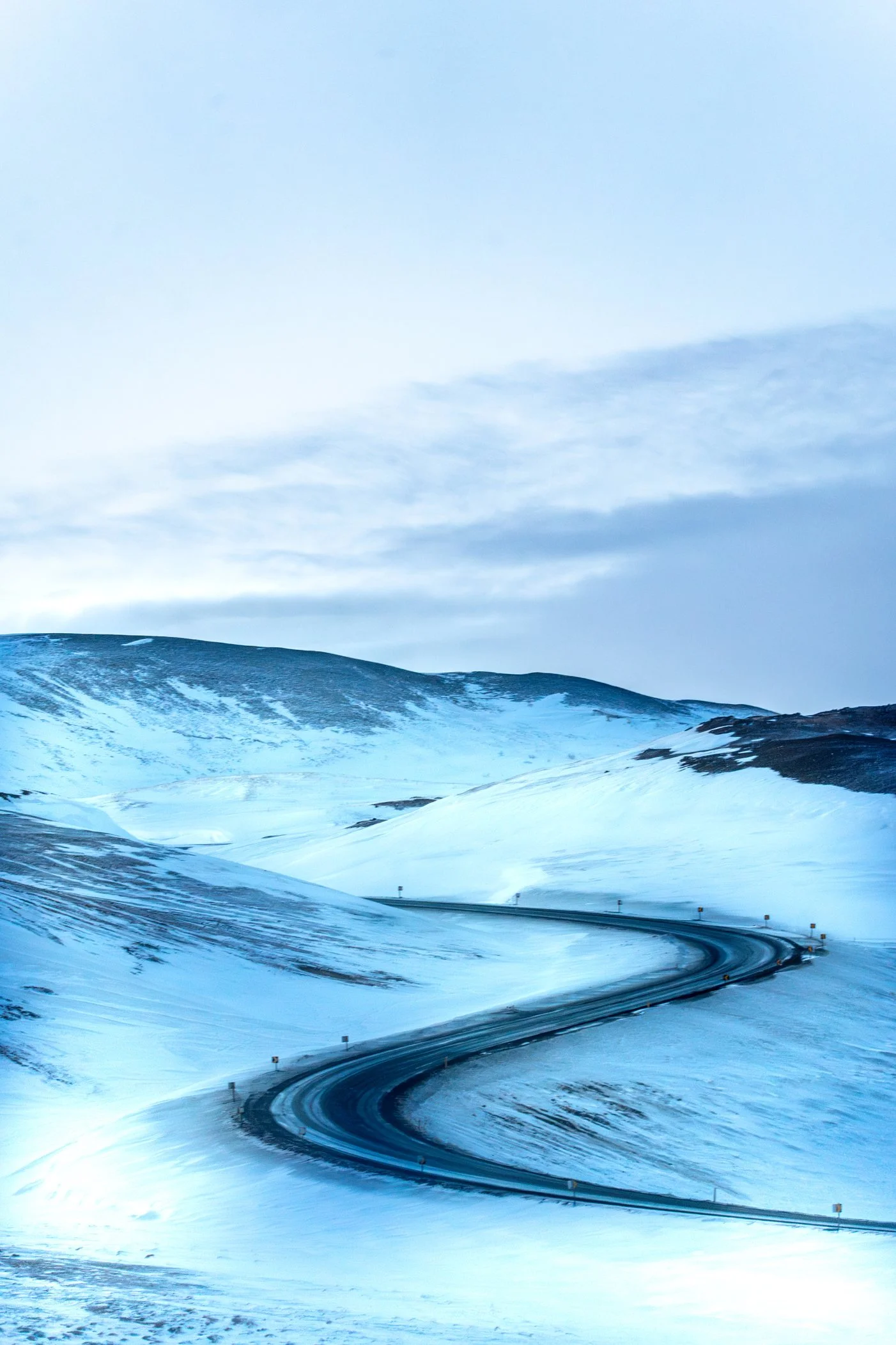 Route serpentant à travers un paysage enneigé avec des montagnes et un ciel partiellement nuageux.