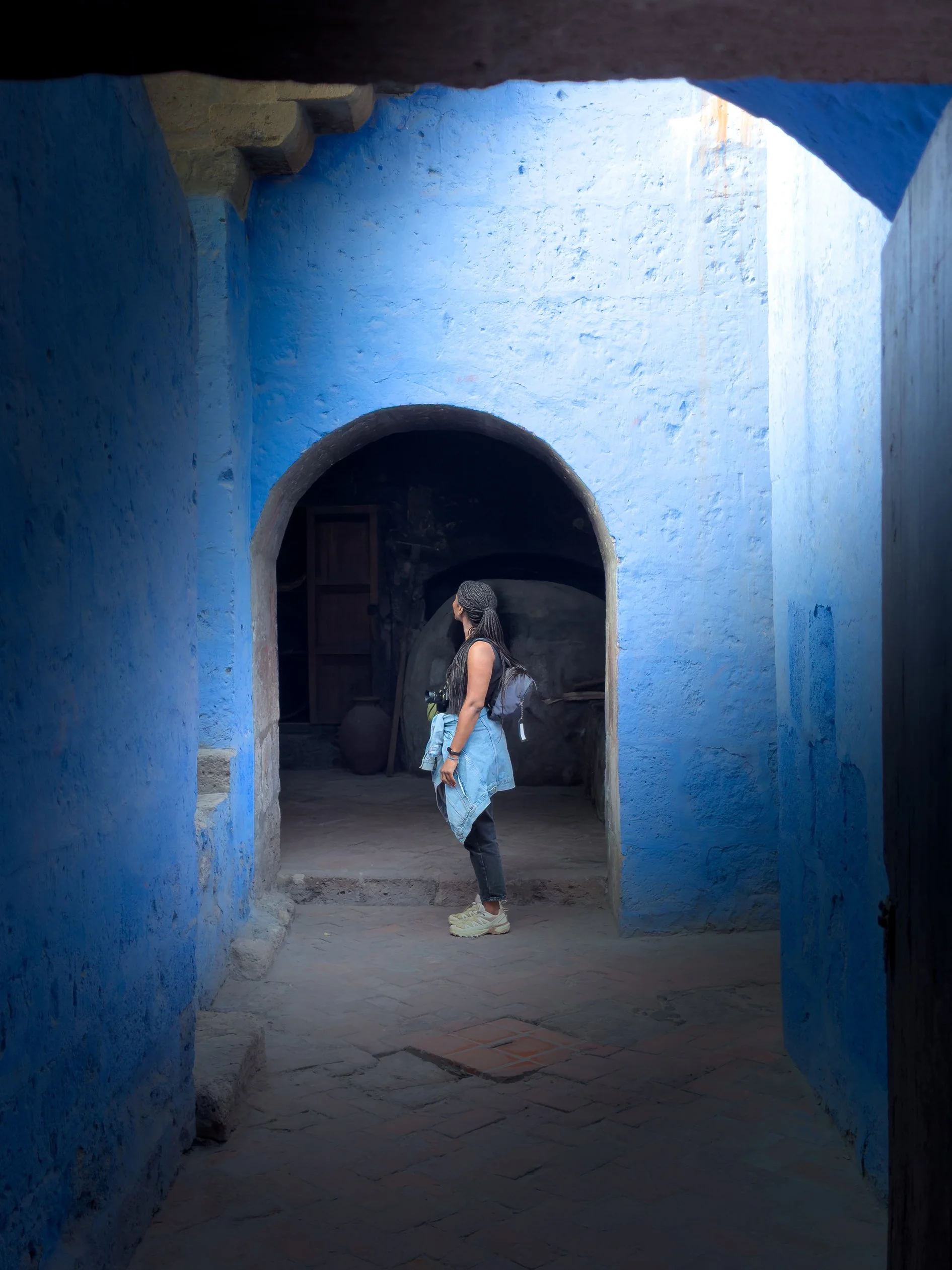 Une personne avec un sac à dos se tient dans un passage voûté au mur bleu dans une ville, regardant de côté.