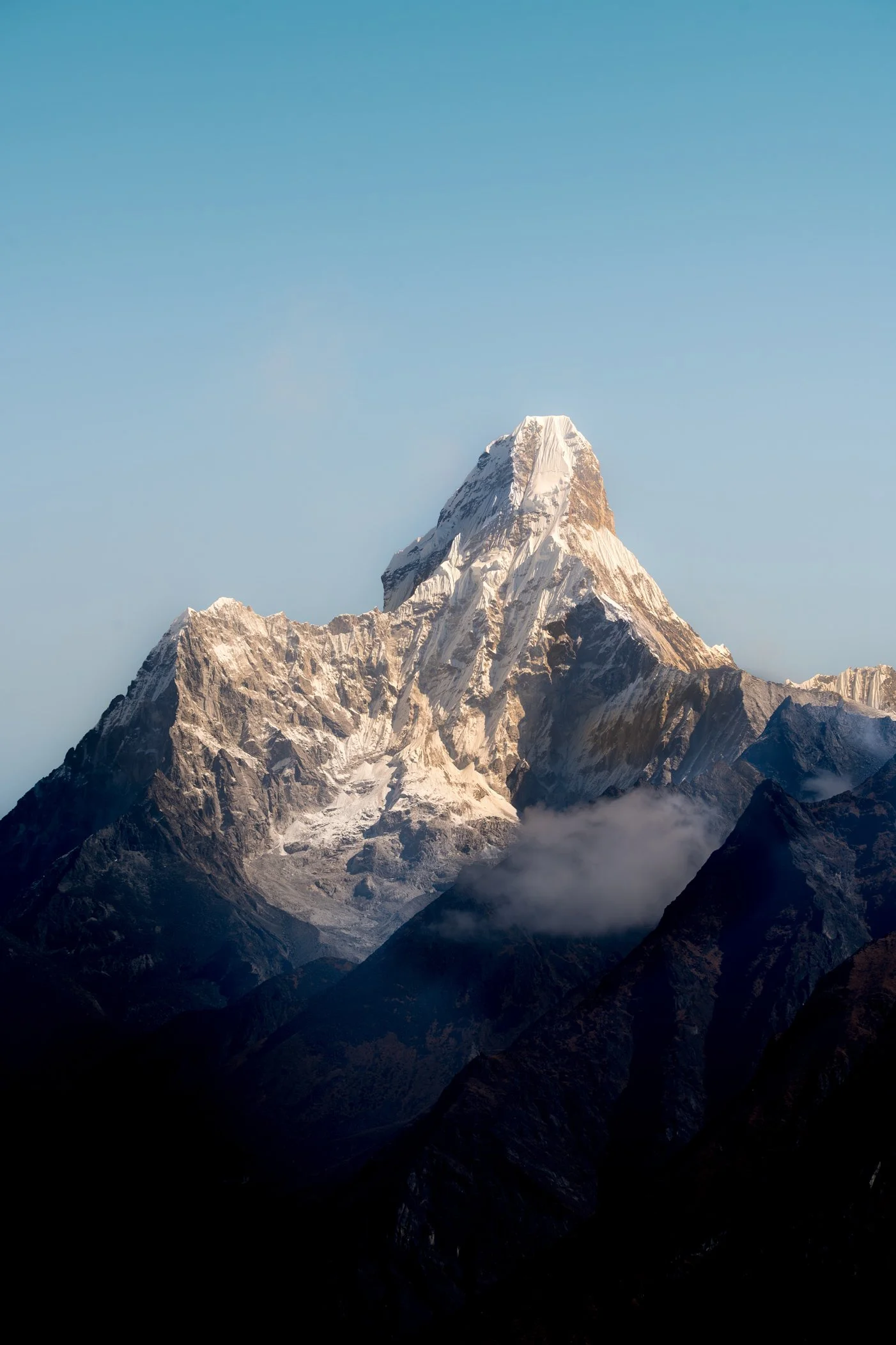 Une montagne enneigée, l'Ama Dablam, avec un ciel bleu clair en arrière-plan et des nuages bas au niveau des sommets.