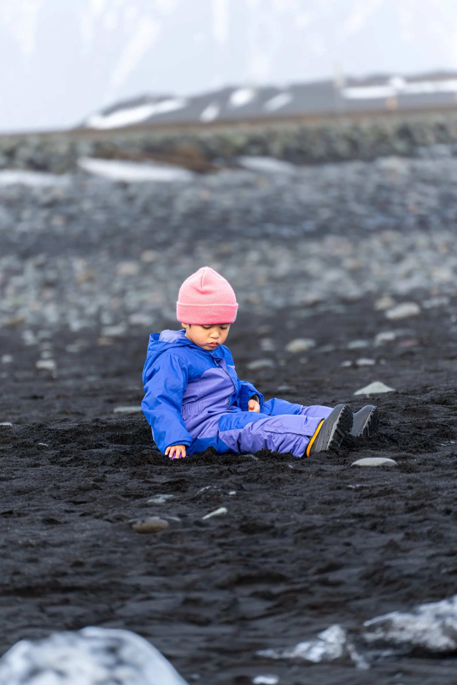 Un enfant assis sur du sable noir, portant une veste bleue, un pantalon violet et un bonnet rose.