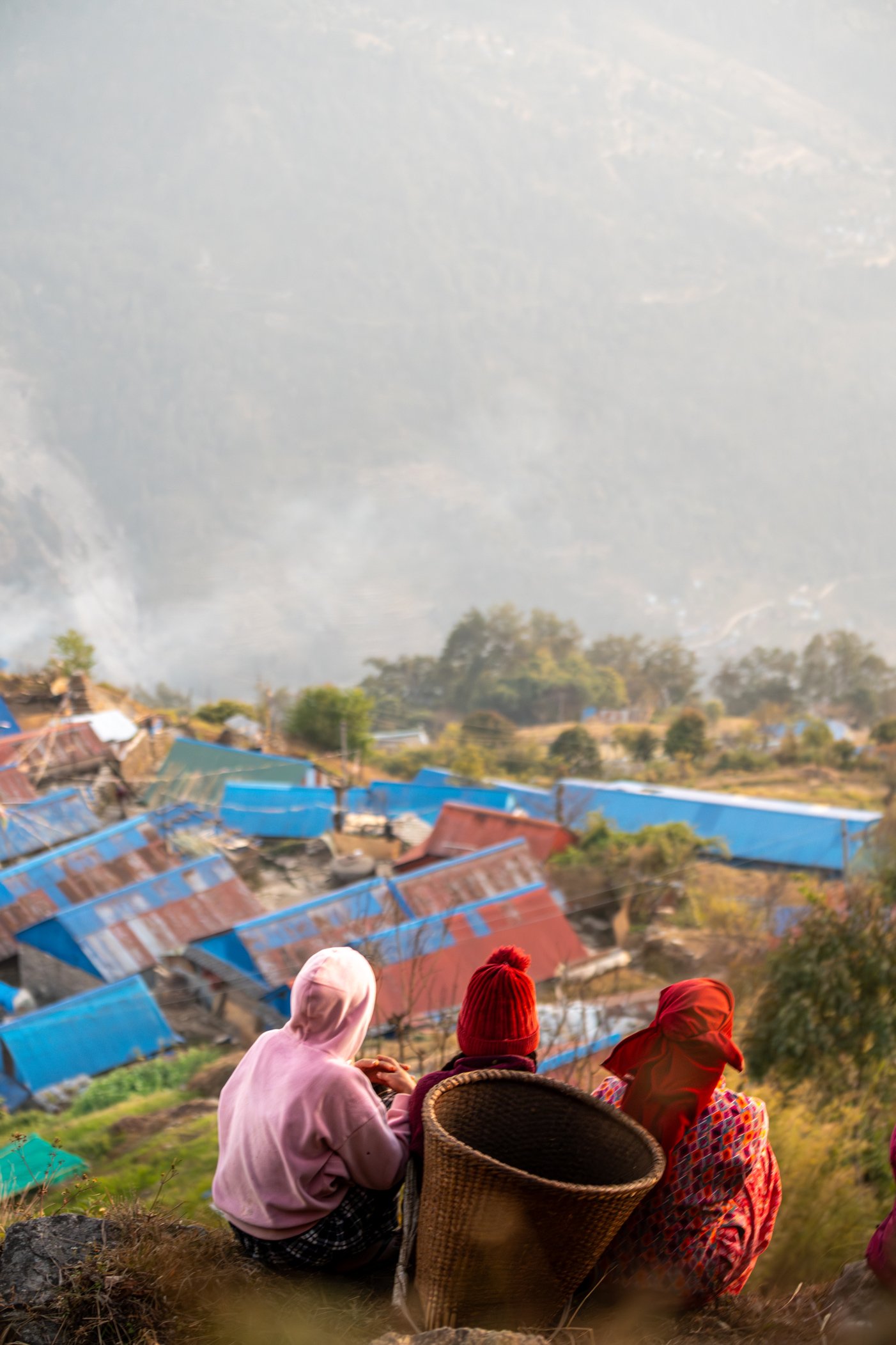 Trois femmes assises sur une colline, regardant un village avec des toits en tôle bleue et rouges, entouré de végétation et de collines.