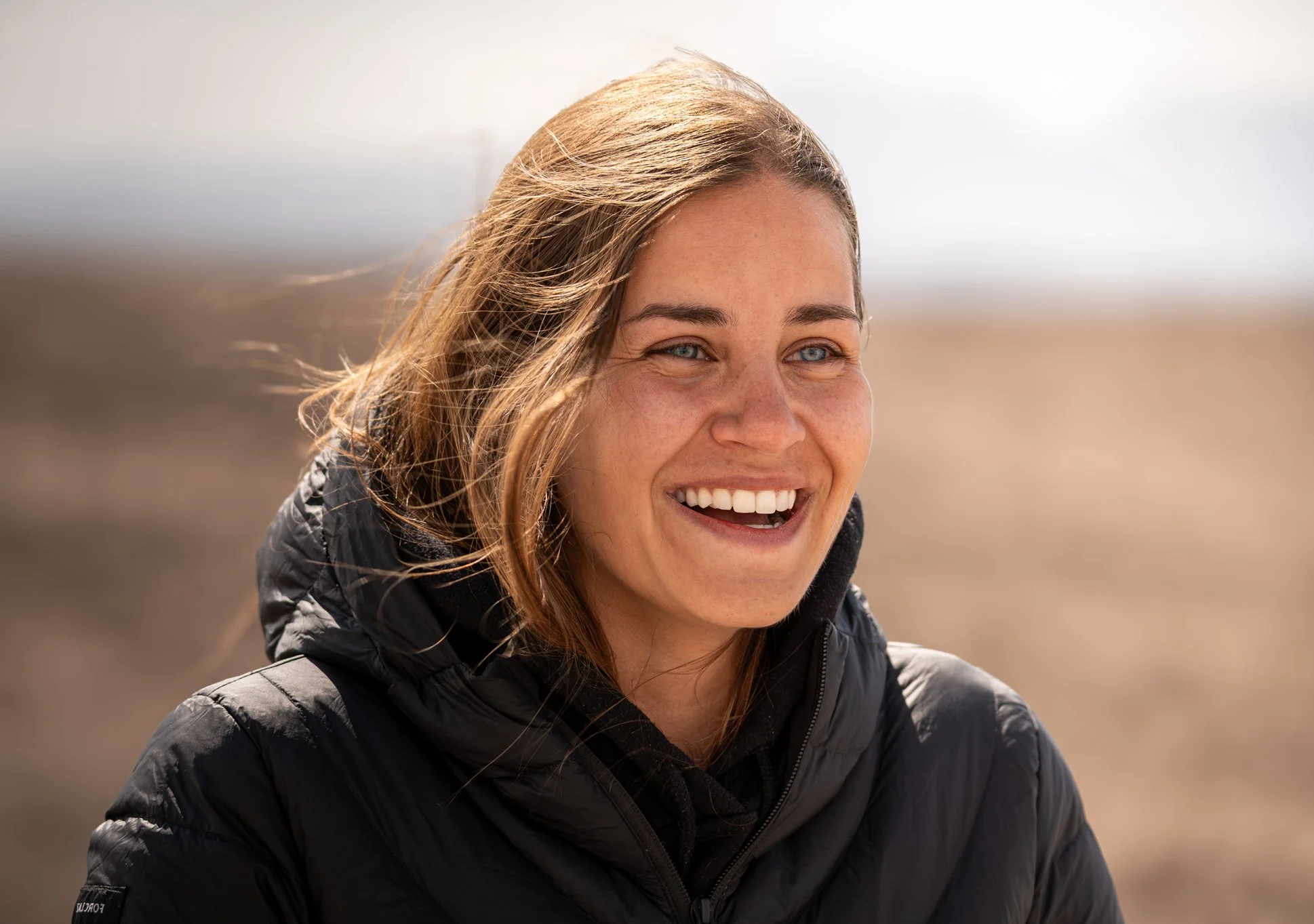 Femme souriante avec des cheveux bruns et des yeux bleus, portant une veste noire, en plein air dans un paysage désertique.