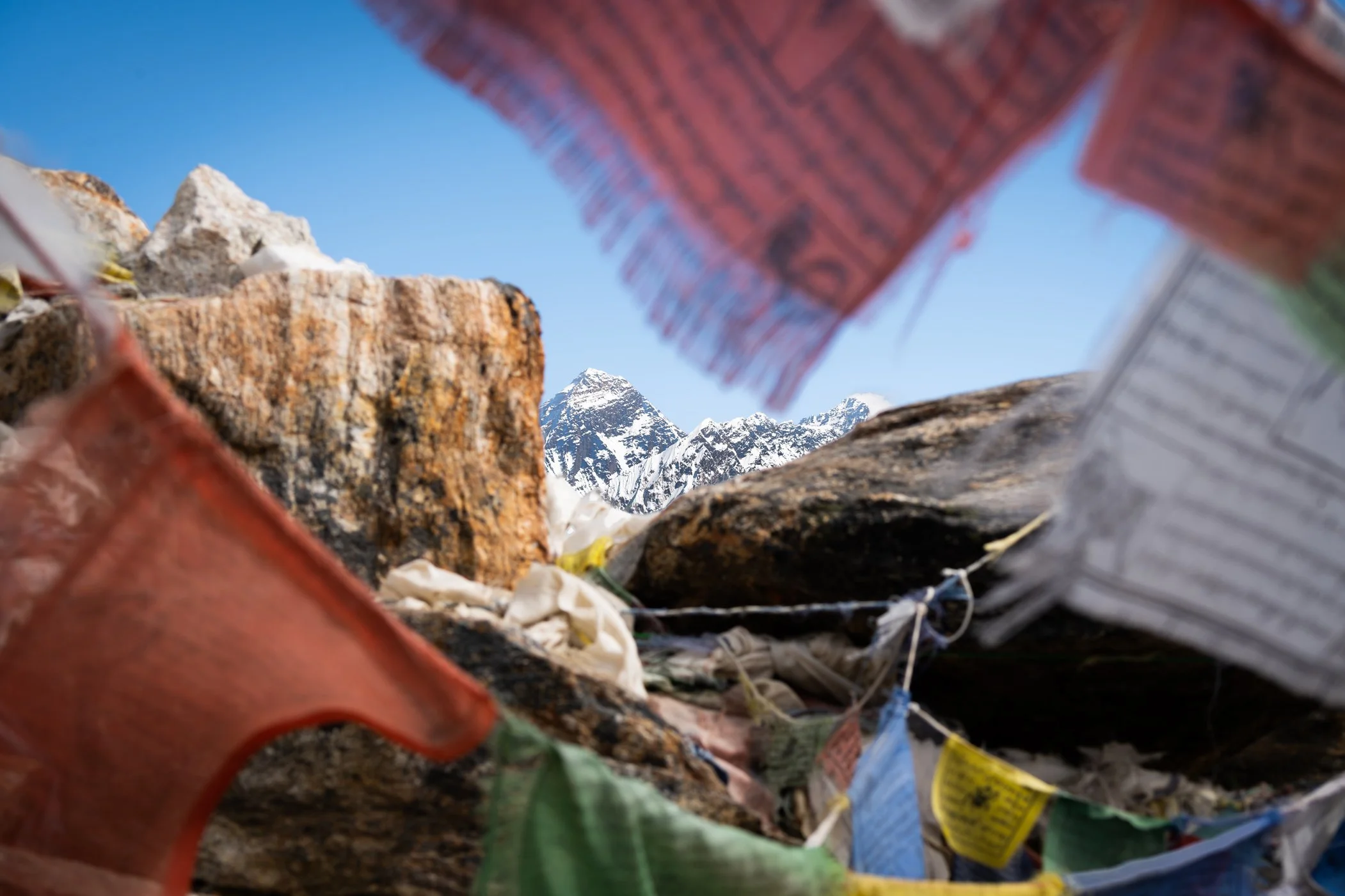 Drapeaux colorés en premier plan, autour de rochers avec sommet enneigé en arrière-plan et ciel bleu.