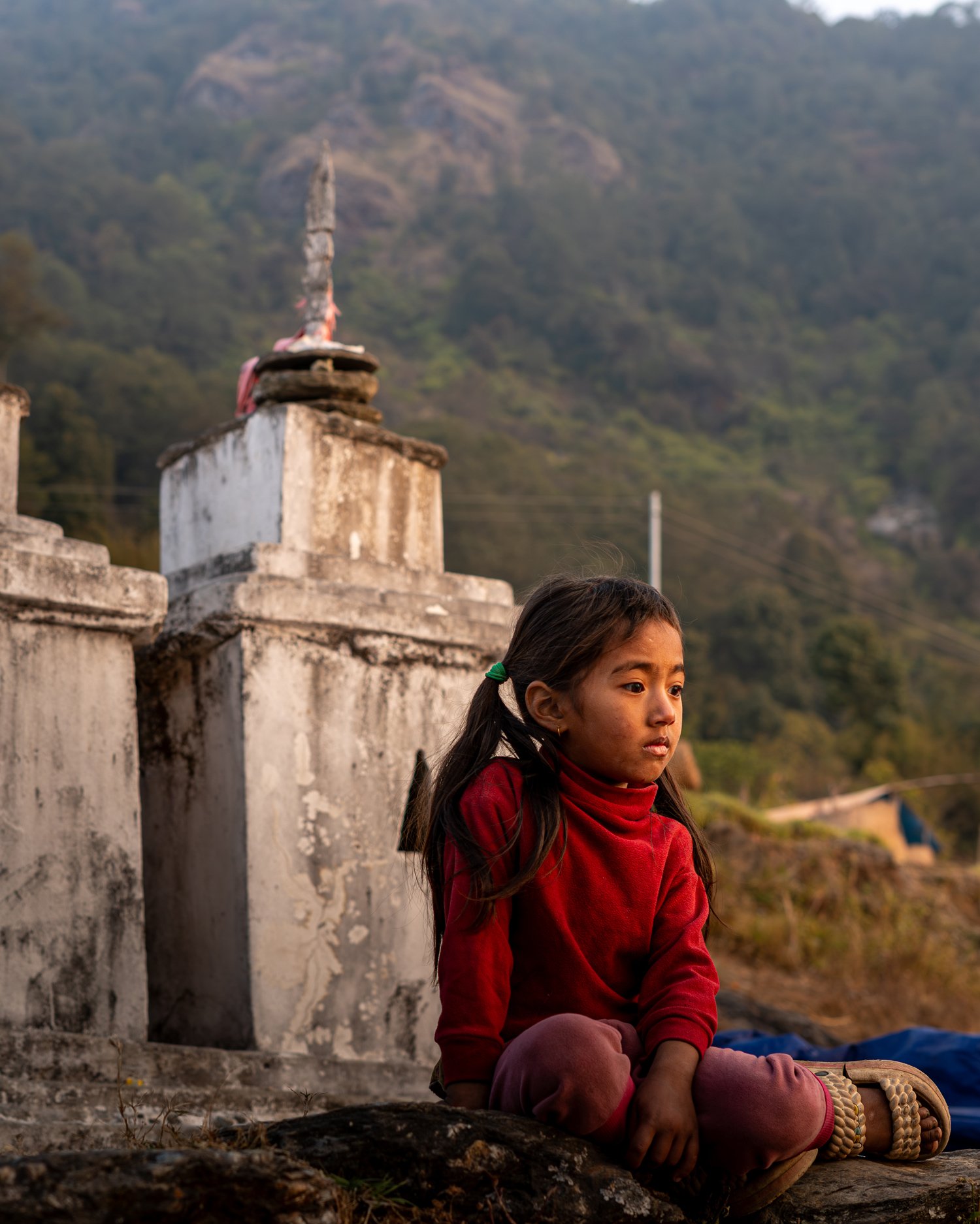 Une jeune fille assise sur le sol dans un paysage rural avec des structures en pierre et une montagne en arrière-plan, portant un vêtement rouge et des bracelets.
