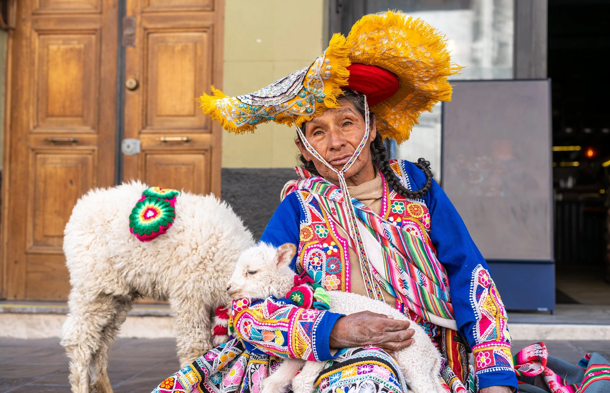 Une femme âgée habillée en costume traditionnel coloré, portant un grand chapeau orné de plumes jaunes, assise avec un agneau blanc sur ses genoux, devant un bâtiment en bois.