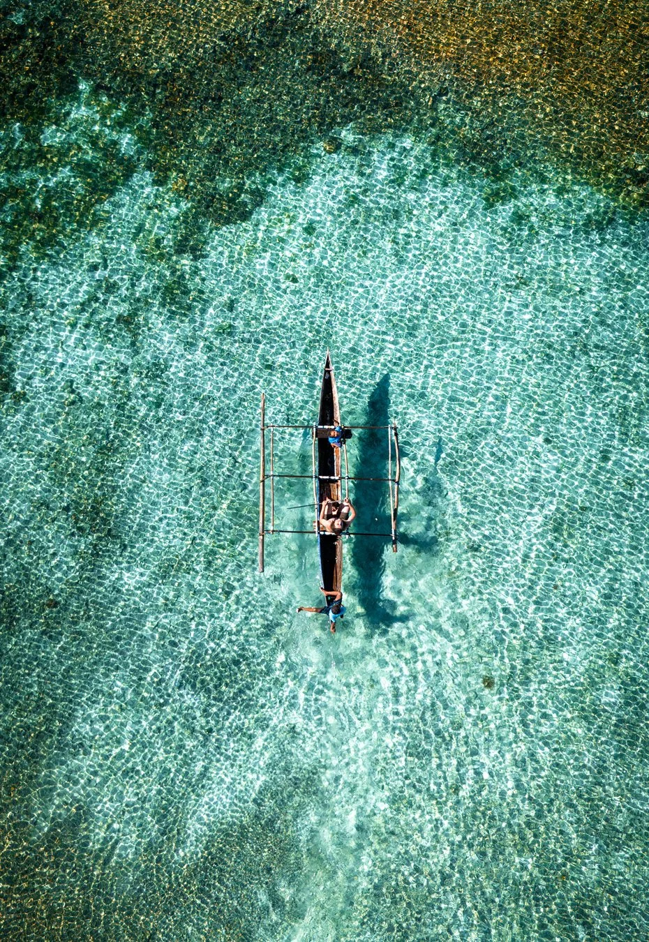 Vue aérienne d'une pirogue en bois dans une mer turquoise claire, avec deux personnes à bord.
