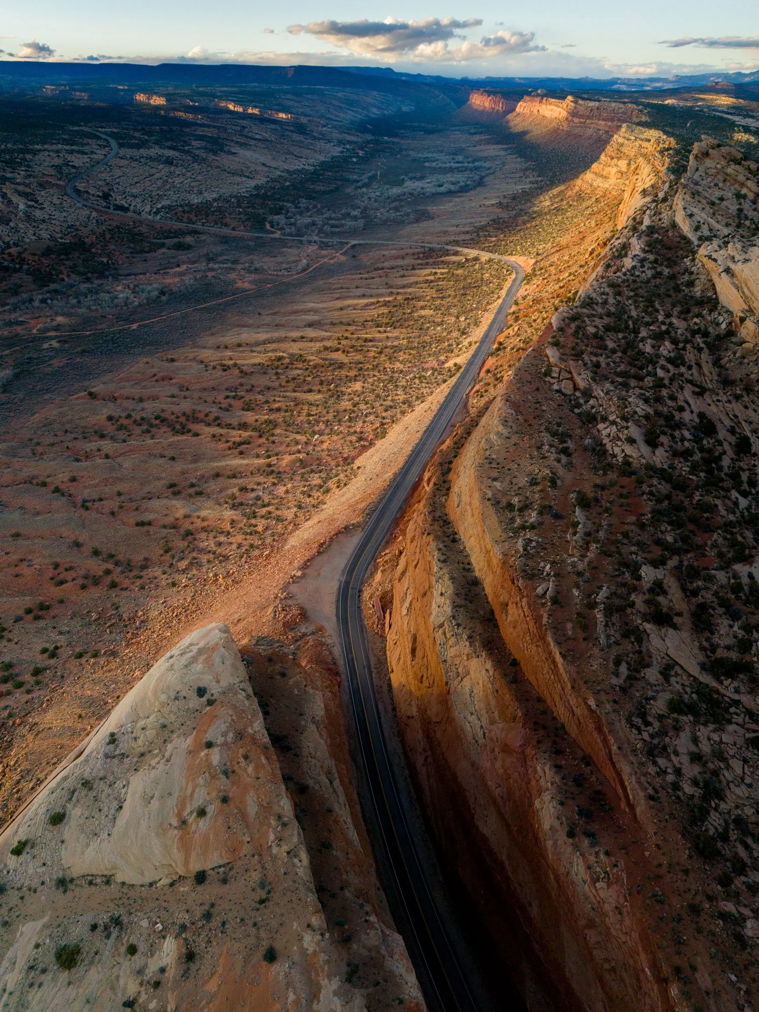 Vue aérienne d'une route sinueuse traversant un canyon désertique avec des formations rocheuses colorées et des montagnes en arrière-plan, sous un ciel nuageux.