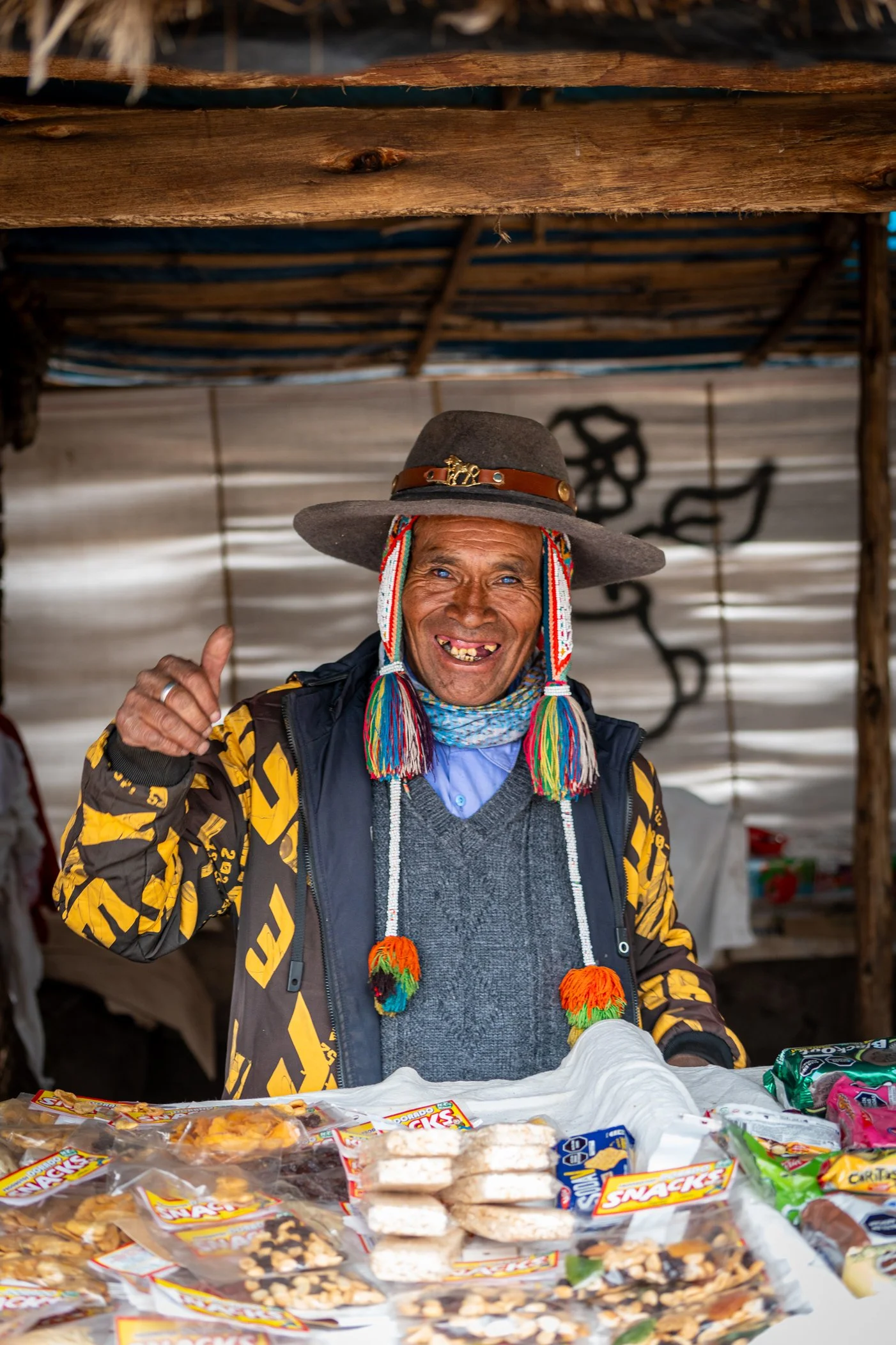 Homme souriant, portant un chapeau folklorique, derrière un stand avec des snacks emballés, en intérieur avec structure en bois.