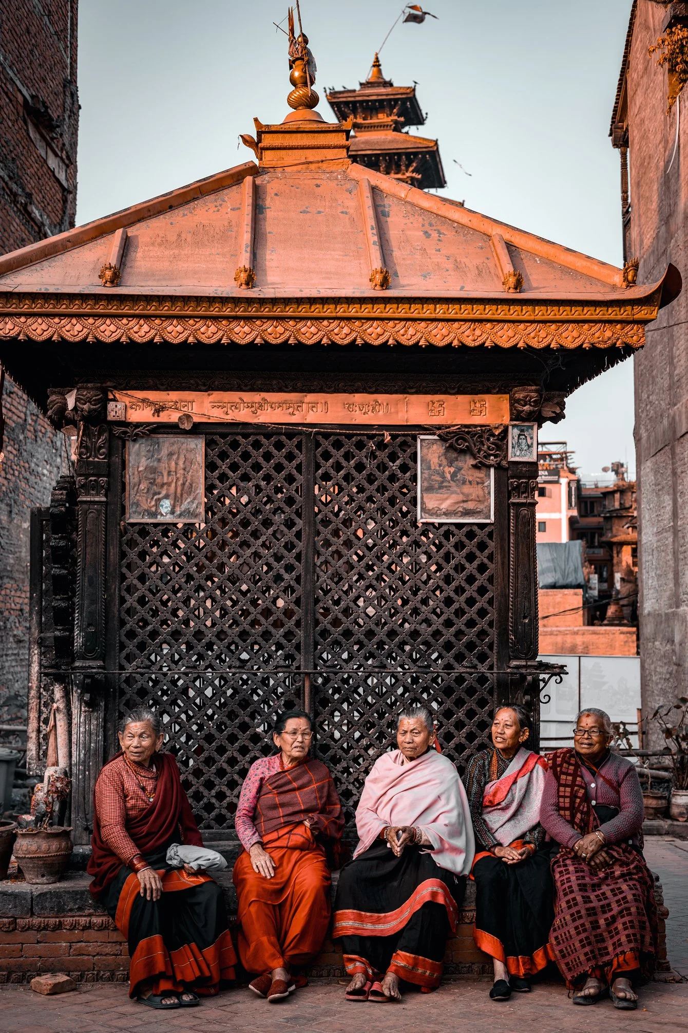 Cinq femmes âgées assises devant un petit pavillon en bois dans une rue de Kathmandu, Népal, avec un temple en arrière-plan.