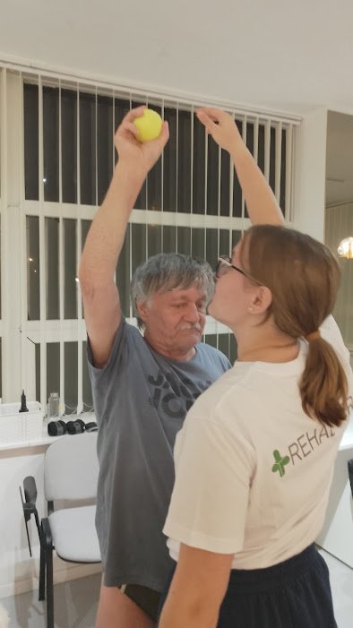 A woman in a white t-shirt with 'REHAB' on the sleeve is standing close to an older man, holding a yellow tennis ball with one hand while they appear to be playing or interacting indoors near a window with vertical blinds.