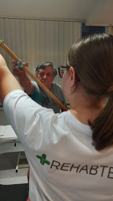 A woman holding a broom or mop handle, with a man in the background looking at her, in a room with vertical blinds.