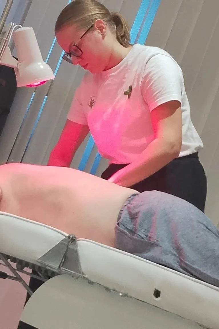 A person lies face down on a massage table with a white pillow under their head, receiving a back massage from a massage therapist in a professional setting. The therapist is wearing glasses and a white shirt, focusing on the person's back.