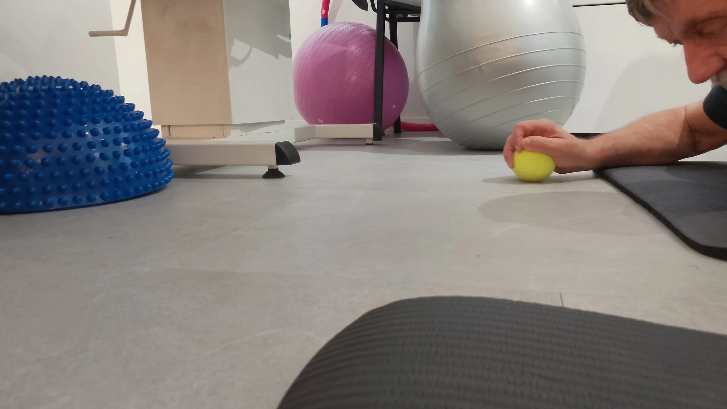 Person exercising on a black yoga mat with a yellow exercise ball, blue textured balance disc, pink and grey exercise balls in the background.