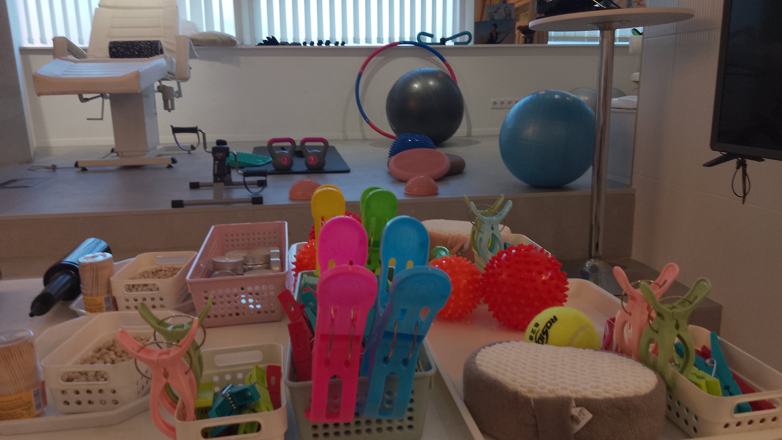A room with therapy and exercise equipment and various toys and supplies, including colorful plastic chairs, textured balls, a tennis ball, and foam sponges, set up for physical therapy or occupational therapy.