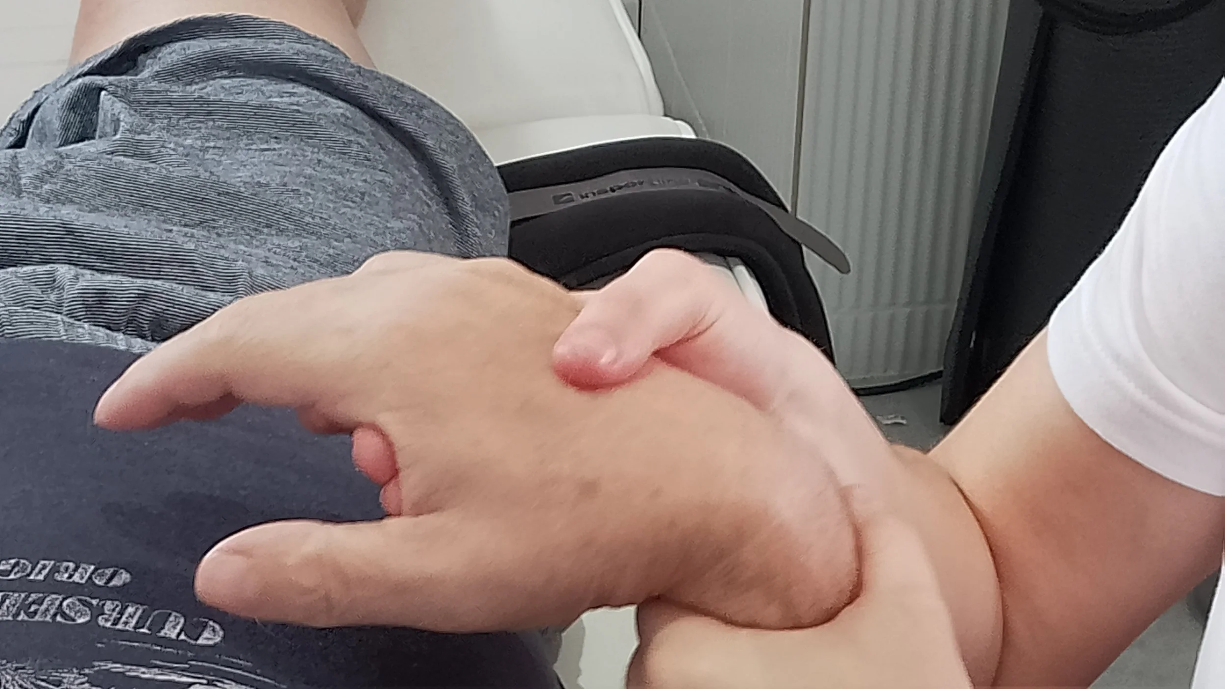 A person receiving a handshake from another individual, with the hand in focus. The background shows part of a gray seat and an acrylic partition.