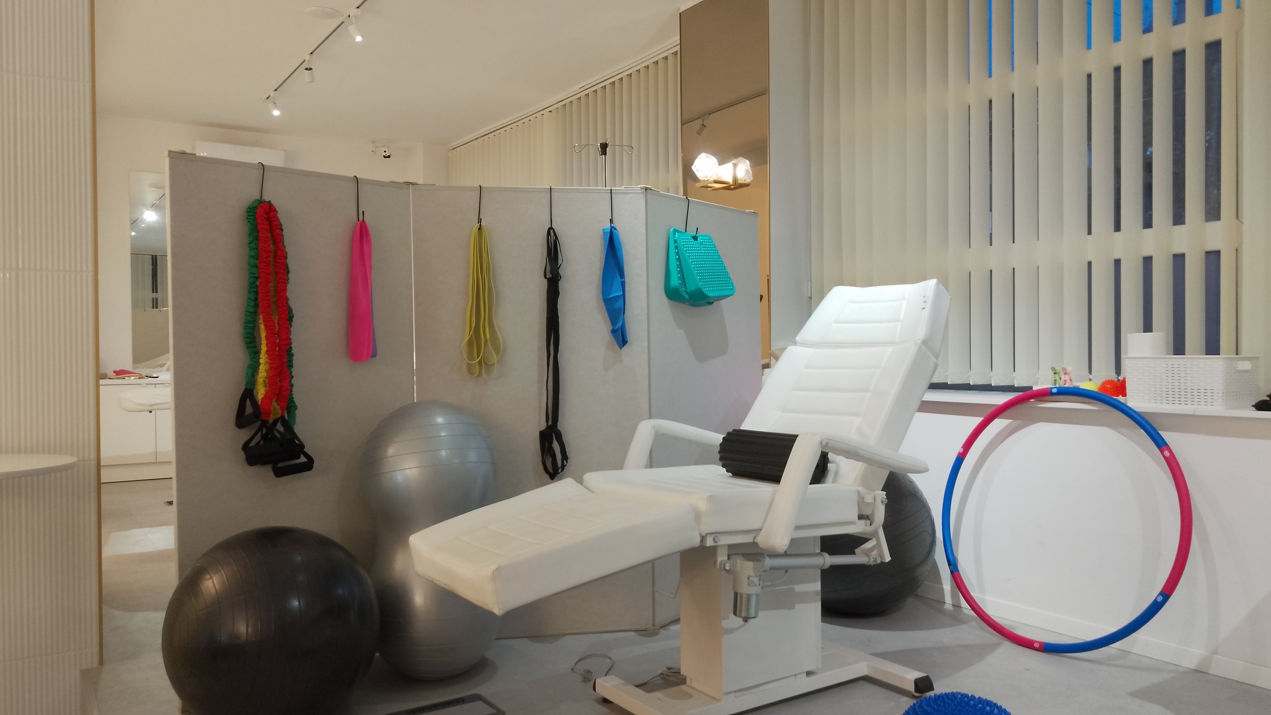 A medical or therapy room with exercise equipment, including shelves with resistance bands and jump ropes, stability balls, a medical chair, and a colorful hula hoop.