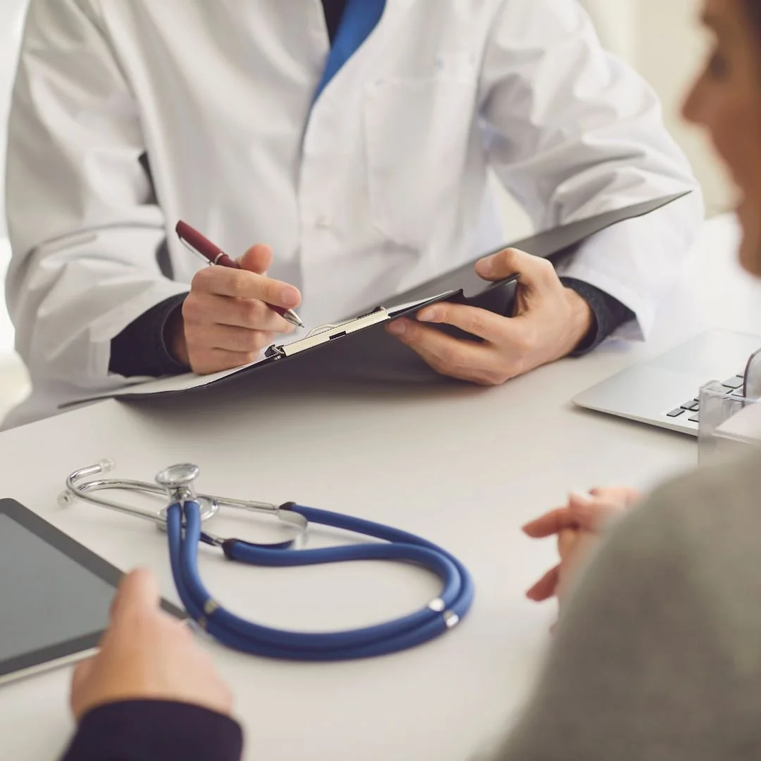 Doctor in a white coat taking notes on a clipboard during a medical consultation, with a stethoscope and laptop on the table.