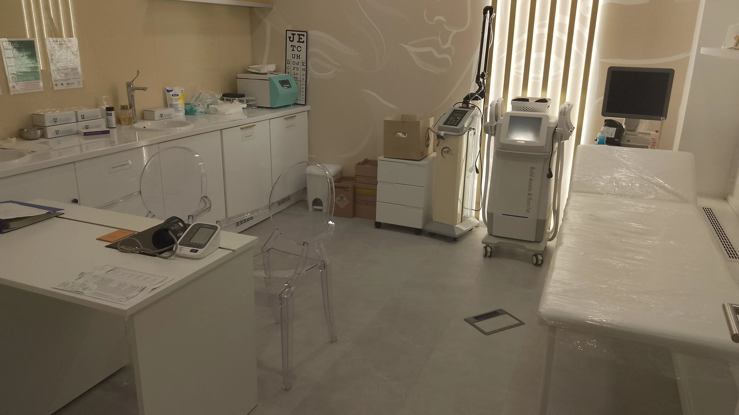 A medical examination room with white cabinets, a bed covered in plastic, medical equipment, and transparent chairs.