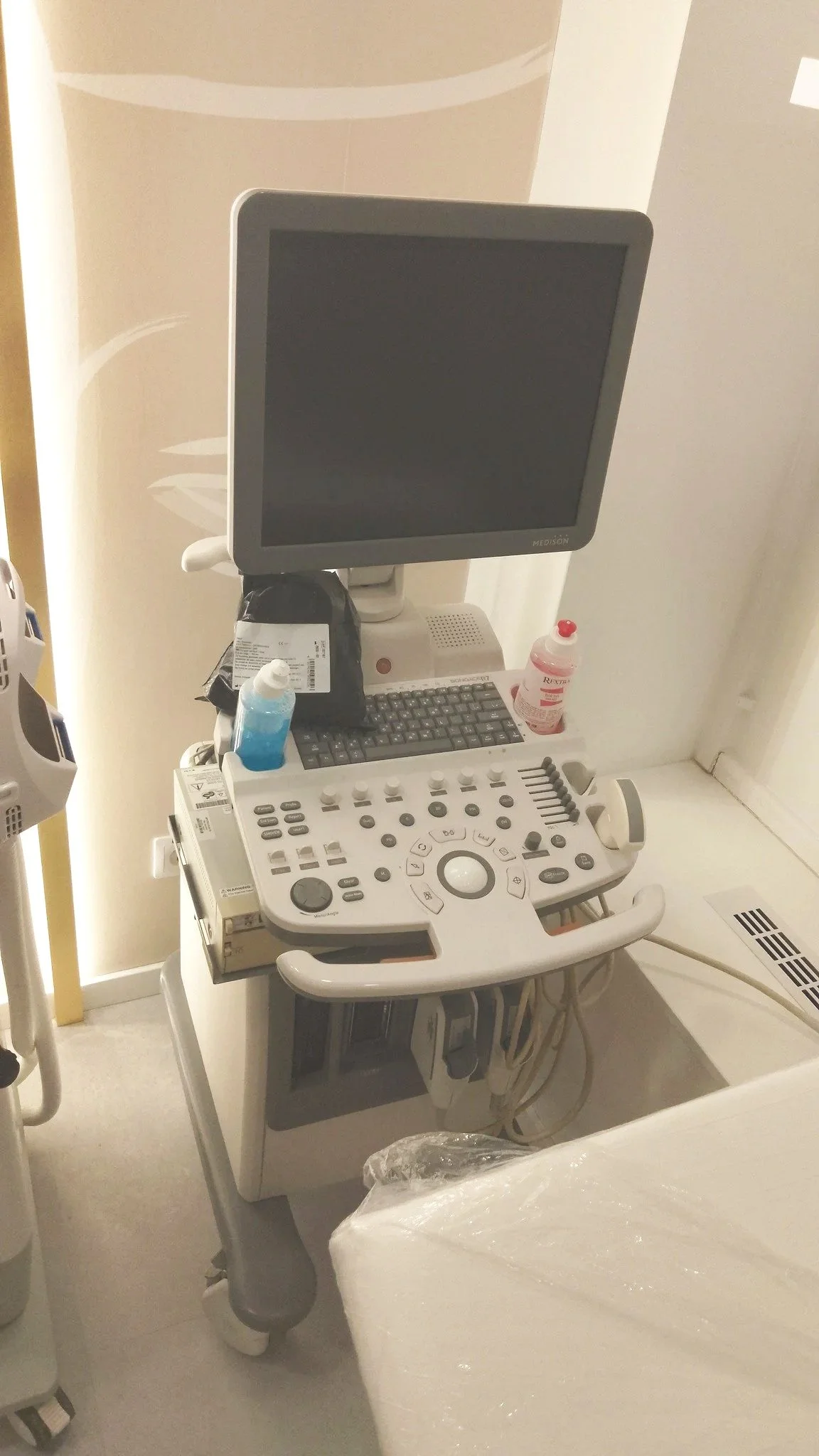 Medical ultrasound machine with a monitor, control panel, and various probes and bottles on top, positioned next to a hospital bed.