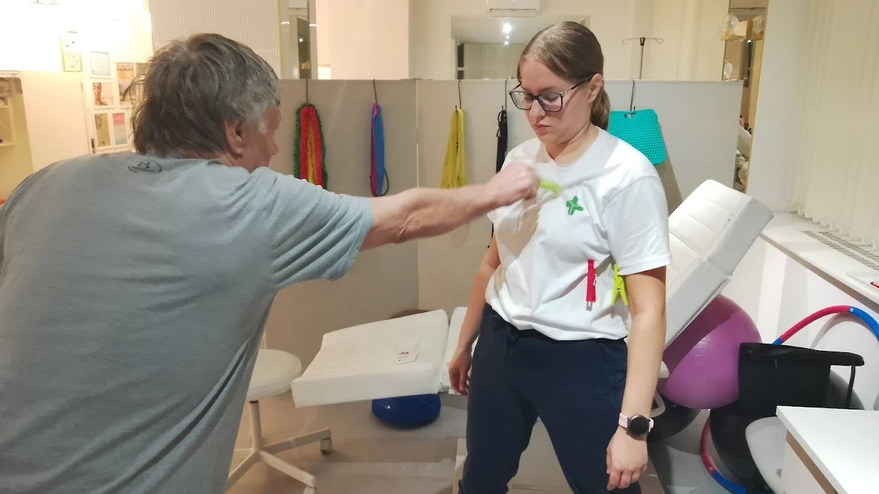 An elderly man is adjusting or touching the shoulder of a young woman in a medical or physiotherapy setting. The woman is standing with a neutral expression, wearing glasses, a white shirt, and navy pants. The background includes medical equipment an