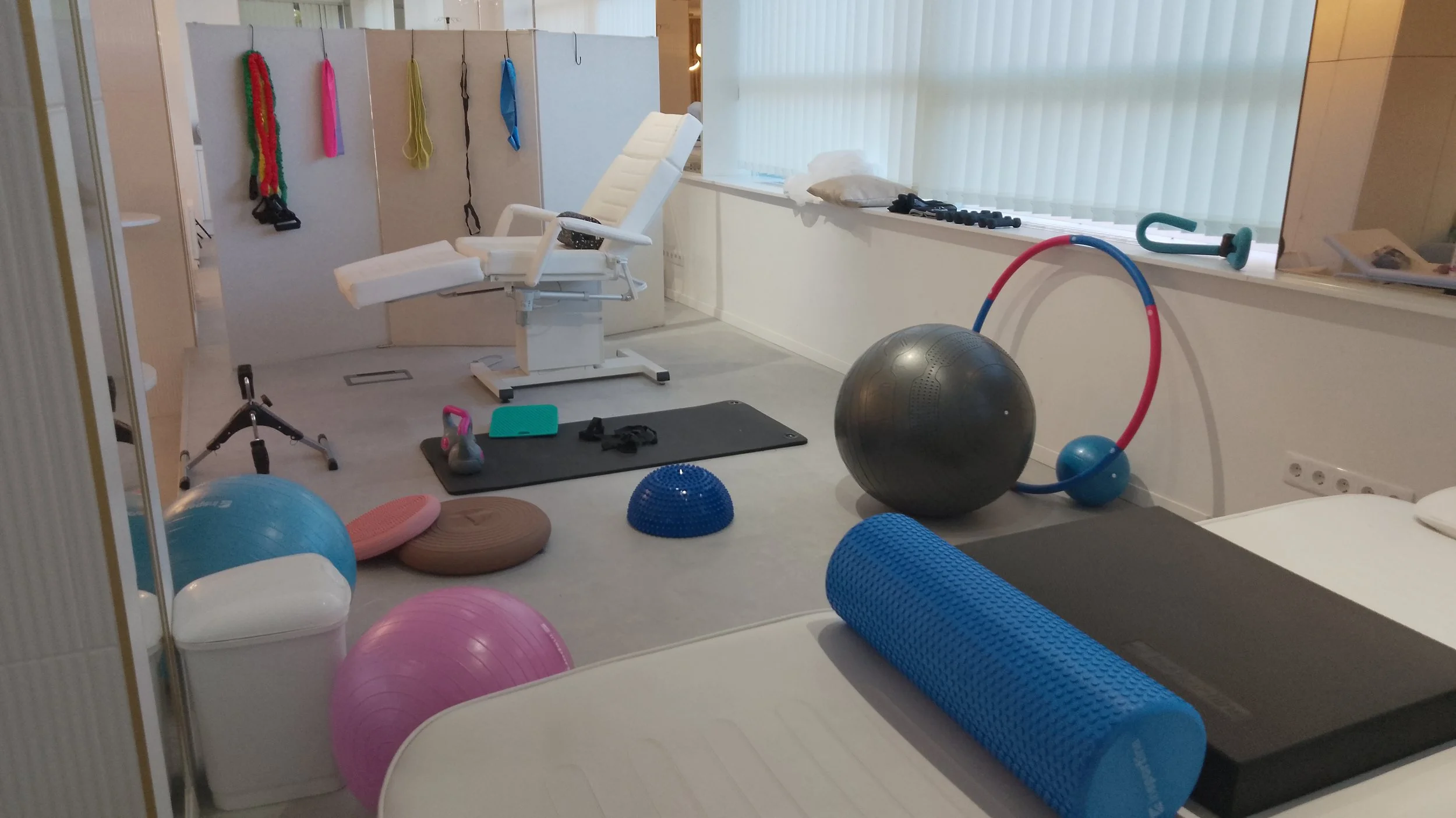 Physical therapy or rehabilitation room with exercise equipment including stability balls, foam rollers, kettlebell, hula hoop, hand weights, and pads, along with a reclined chair and a privacy partition.