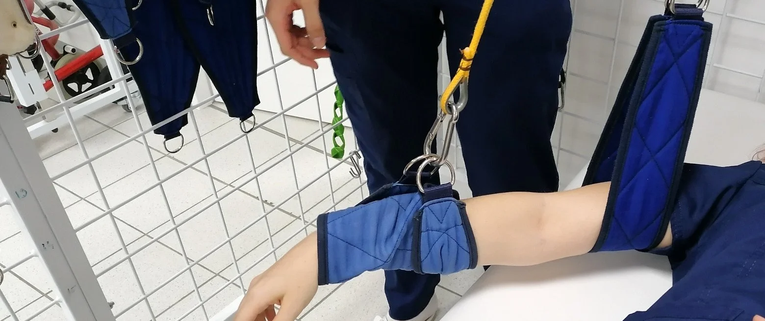 Person receiving physical therapy or rehabilitation on their arm, which is supported by a blue sling, in a medical or clinical setting.