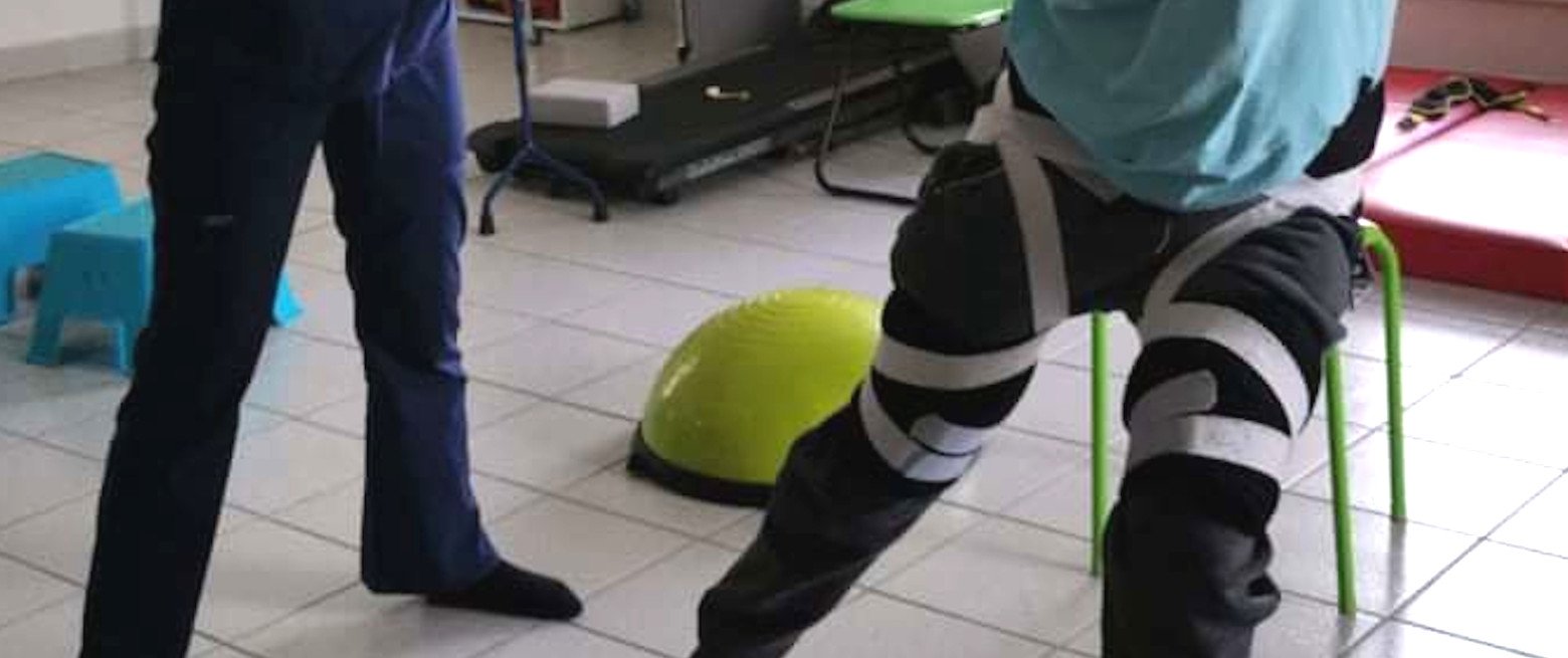 A person sitting on a small green chair with their legs apart, wearing black pants with white stripes, a blue backpack, and a blue shirt. Another person standing nearby is partially visible, dressed in dark pants. The room has a tiled floor, and ther