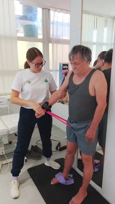 A woman holding a device to a man's arm during a physical therapy session, with the man standing on a mat in a clinical setting.