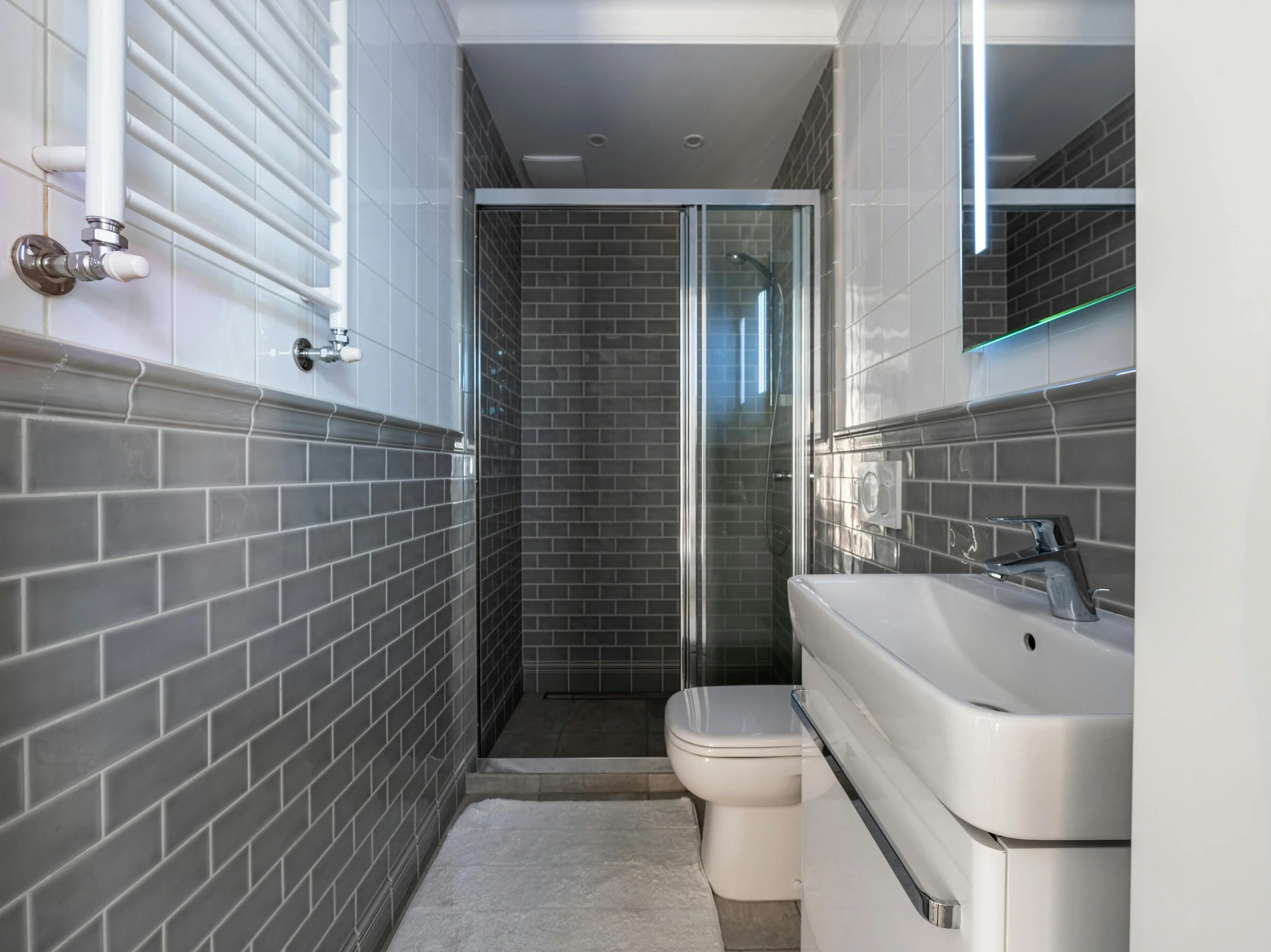 Modern bathroom with gray subway tile walls, glass shower enclosure, white sink, toilet, and a large mirror with LED lighting.