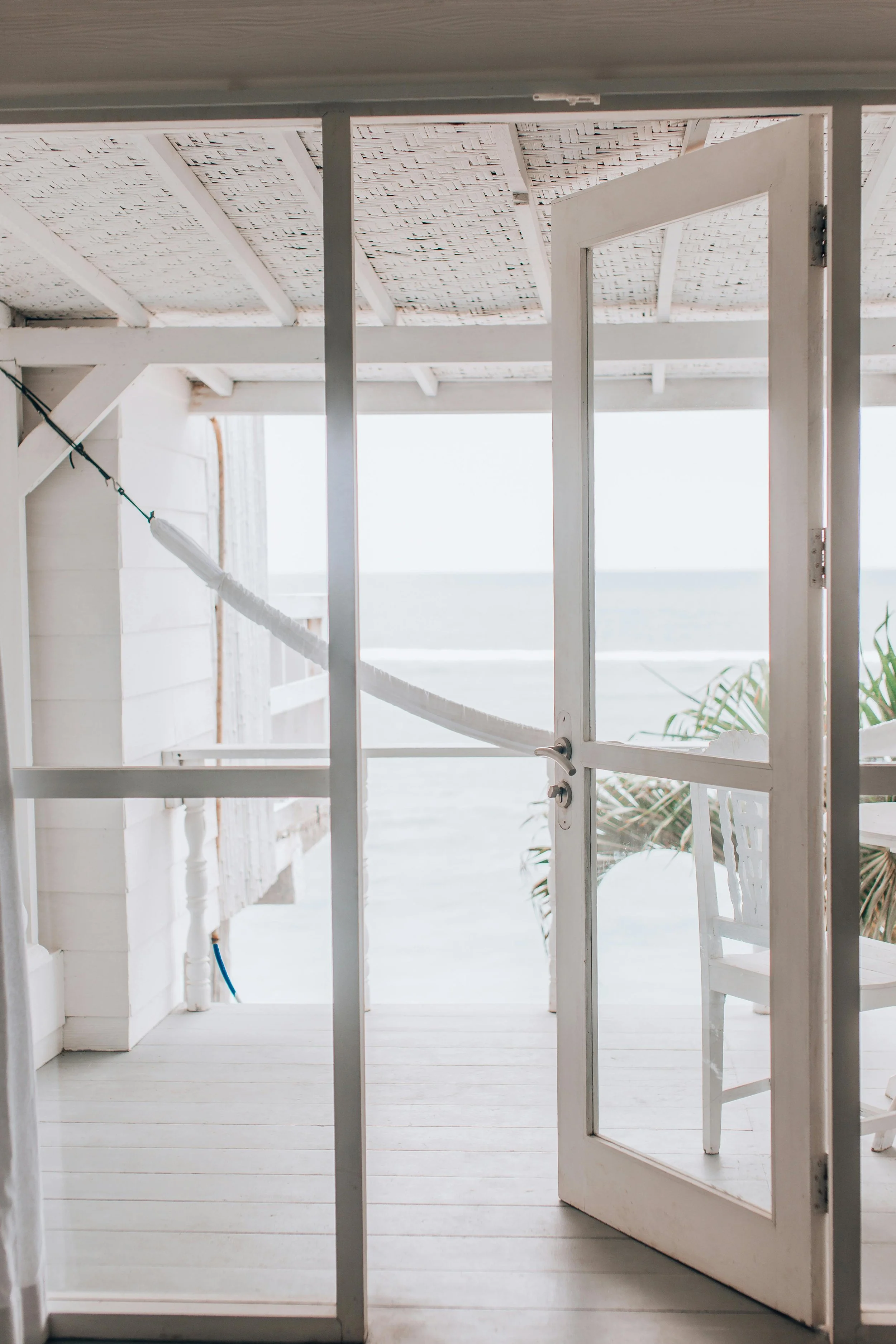 View of a white porch with an open door, overlooking the ocean and partly visible palm leaves.