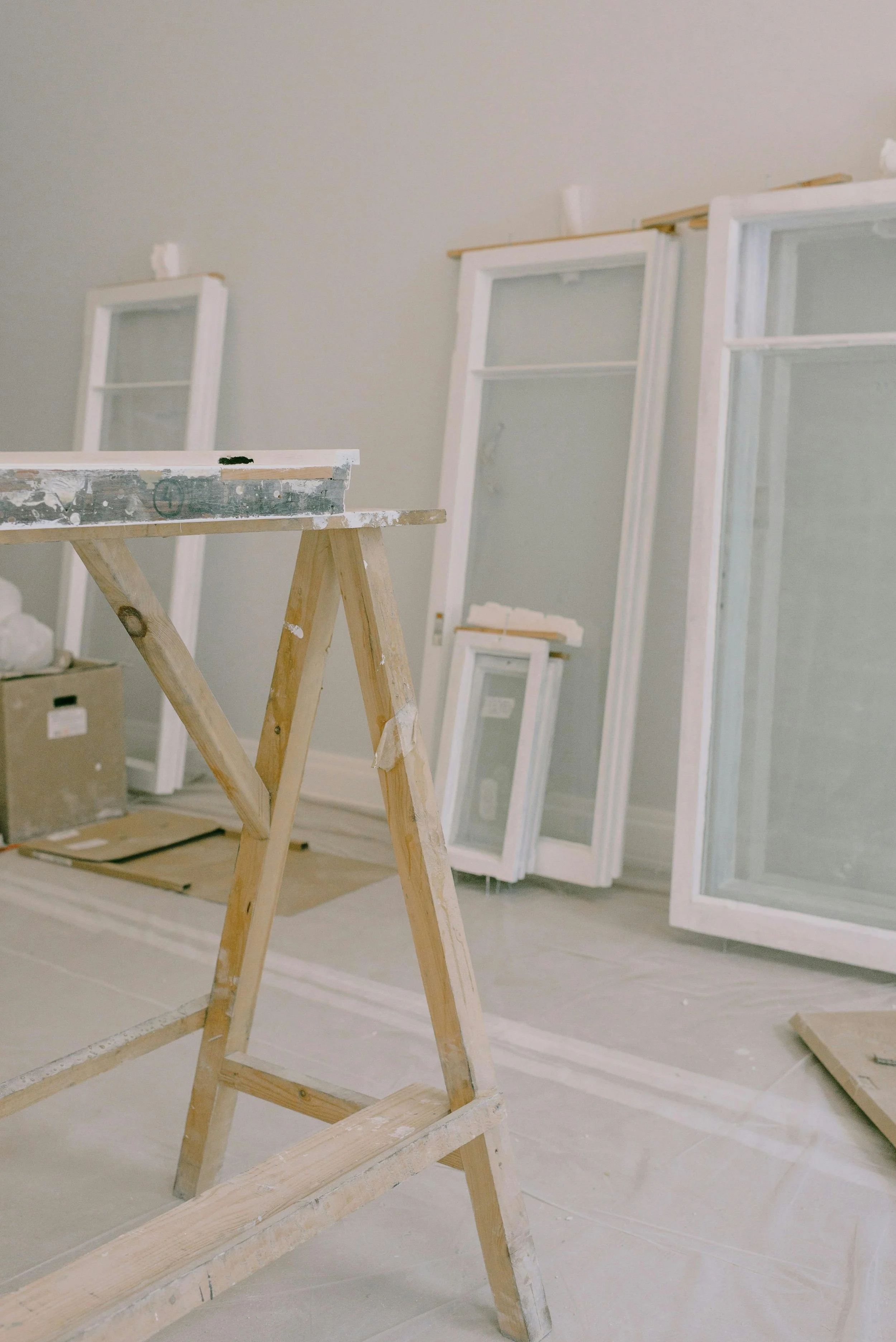 Interior of a room under renovation with multiple white-framed windows and a wooden sawhorse in the foreground.