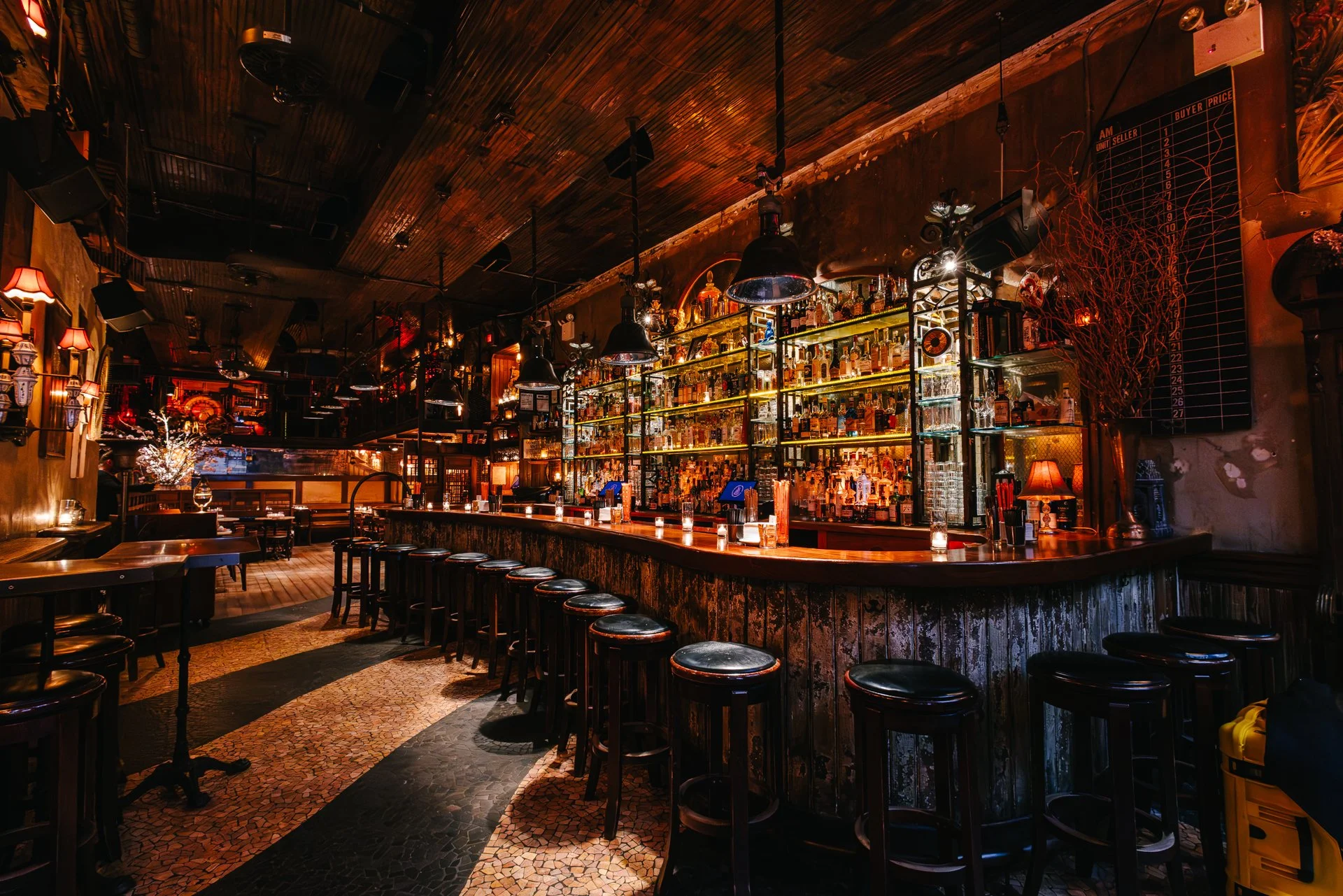 A cozy bar with a wooden counter, high black stools, and illuminated shelves filled with liquor bottles. Warm lighting and vintage decor create an inviting atmosphere.