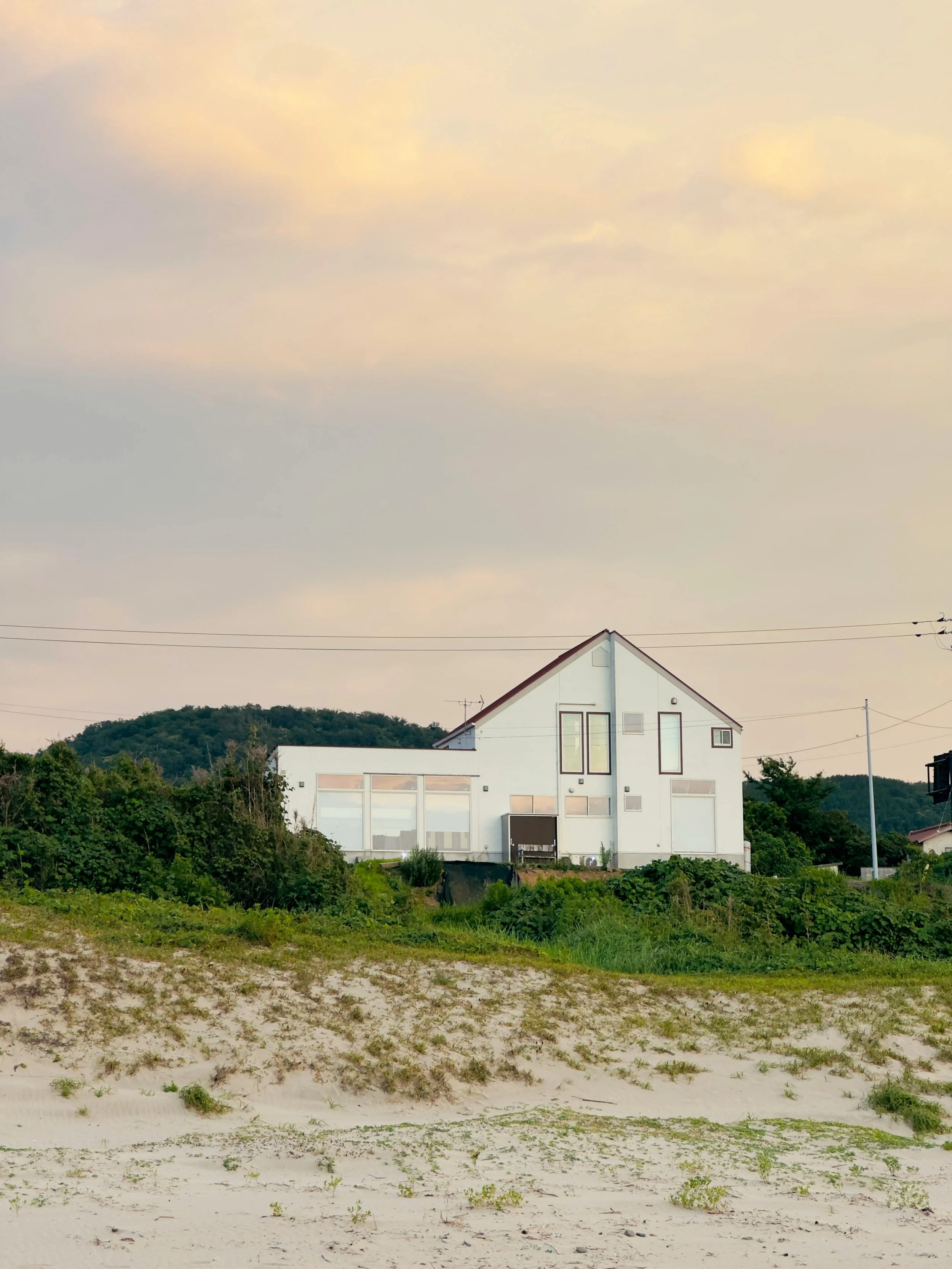 A modern white house on a small hill with grass and sandy terrain in the foreground, hills and a cloudy sky in the background.