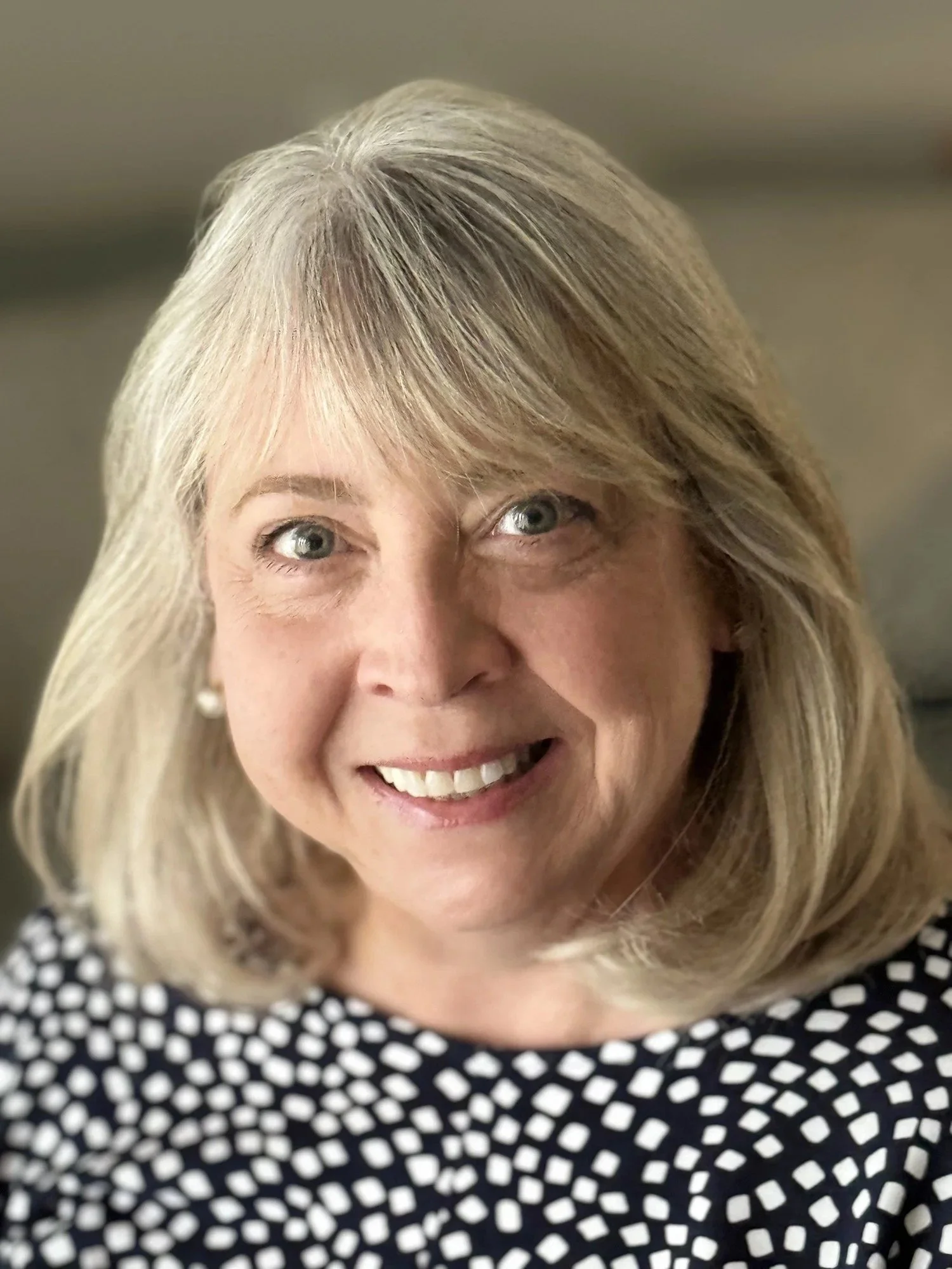 A close-up portrait of a smiling woman with shoulder-length blonde hair, wearing a black and white polka dot top and pearl earrings.