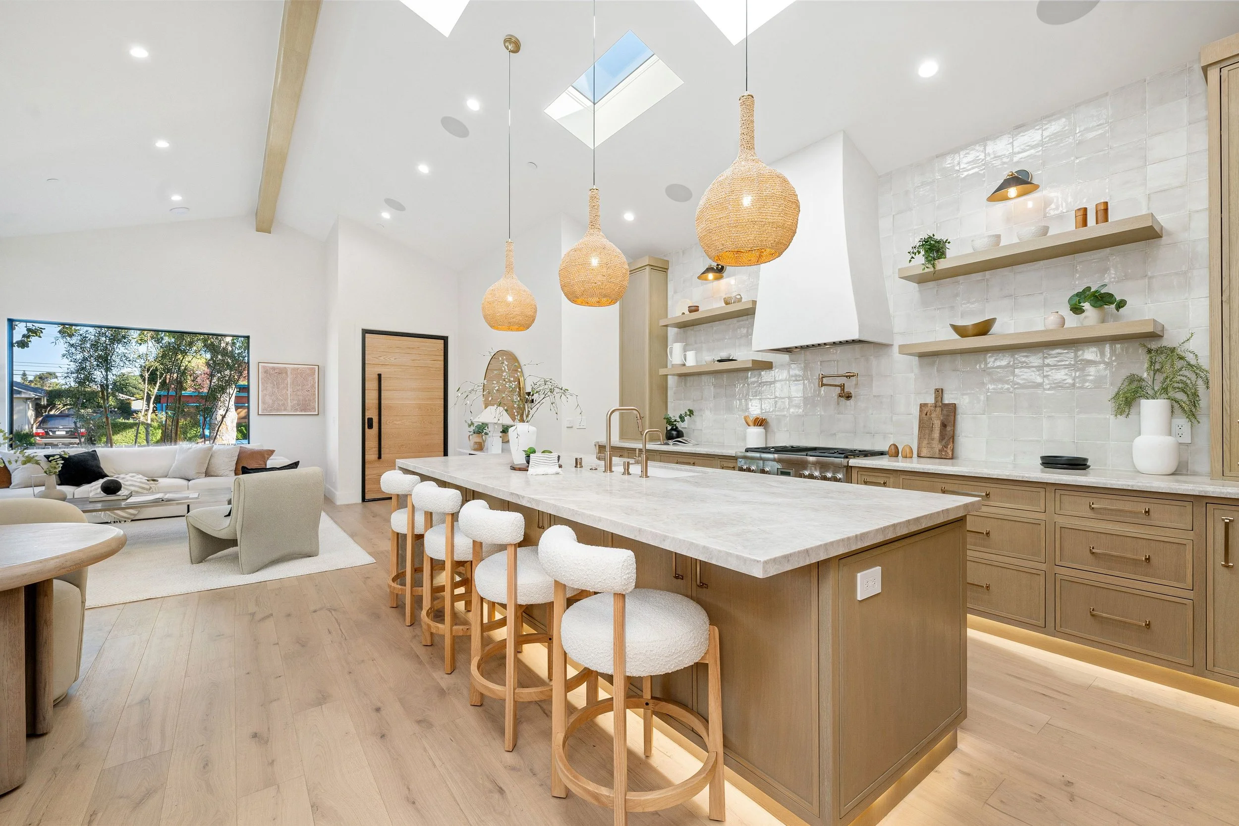 Open-concept kitchen with a large island, four cozy bar stools, pendant lights, and a living area with a beige sofa and window view.