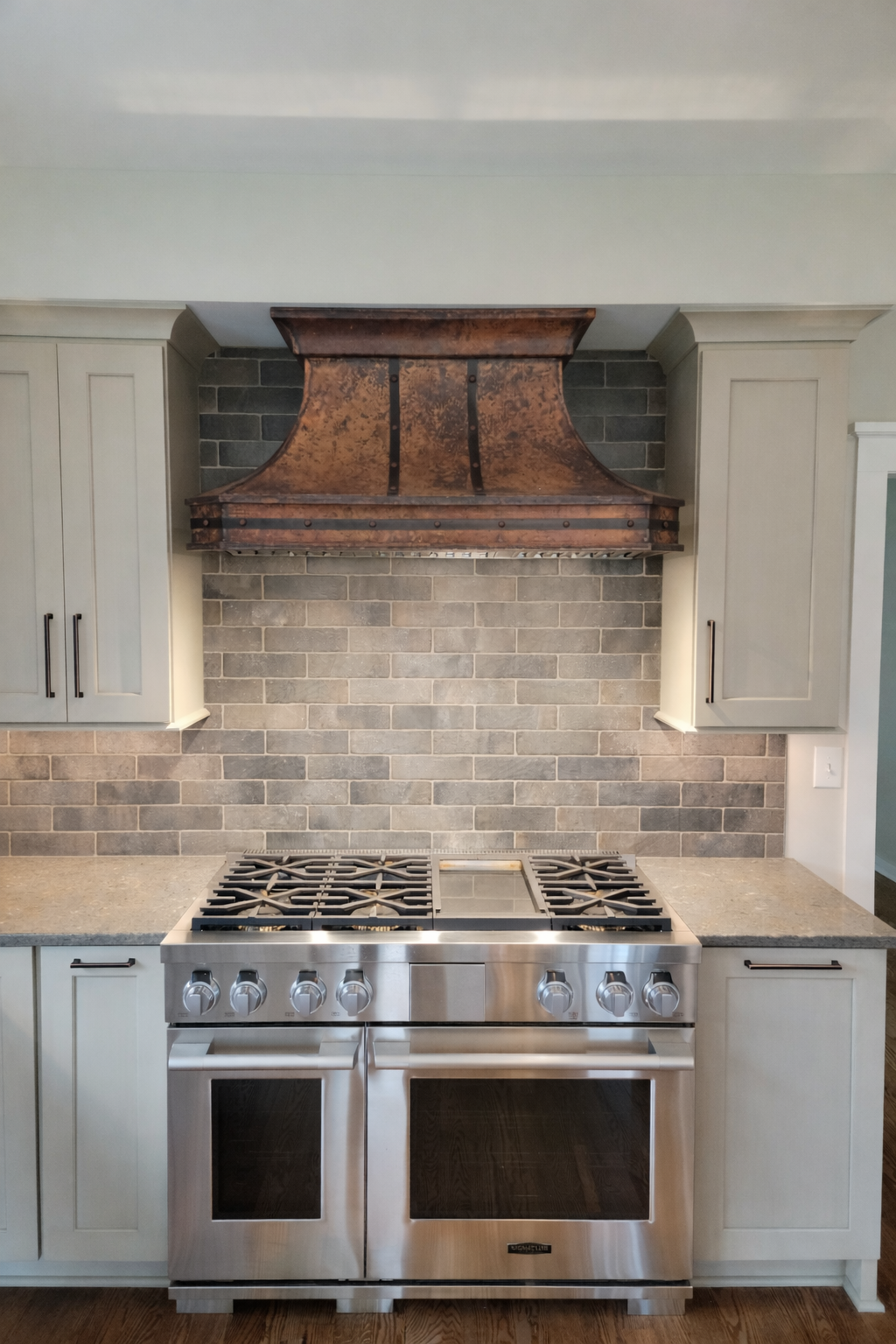 A custom copper range hood is a luxury focal  point in this warm and inviting kitchen. Off white cabinets and textural brick backsplash contribute to the relaxed refinement evident throughout the project. 