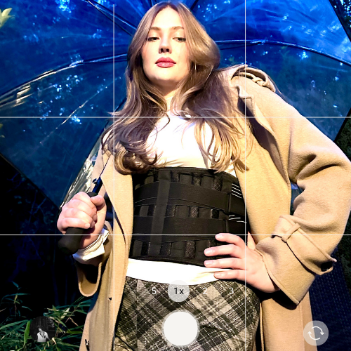 Junge Frau im Freien, trägt einen Mantel und einen Korsett, hält einen Regenschirm, im Hintergrund sind Bäume und blauer Himmel sichtbar.