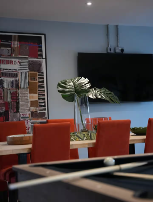 Indoor dining area with a wooden table, orange upholstered chairs, a tall glass vase with large green tropical leaves, and a flat screen TV mounted on the blue wall. There's also an abstract art piece and a foosball table in the foreground.