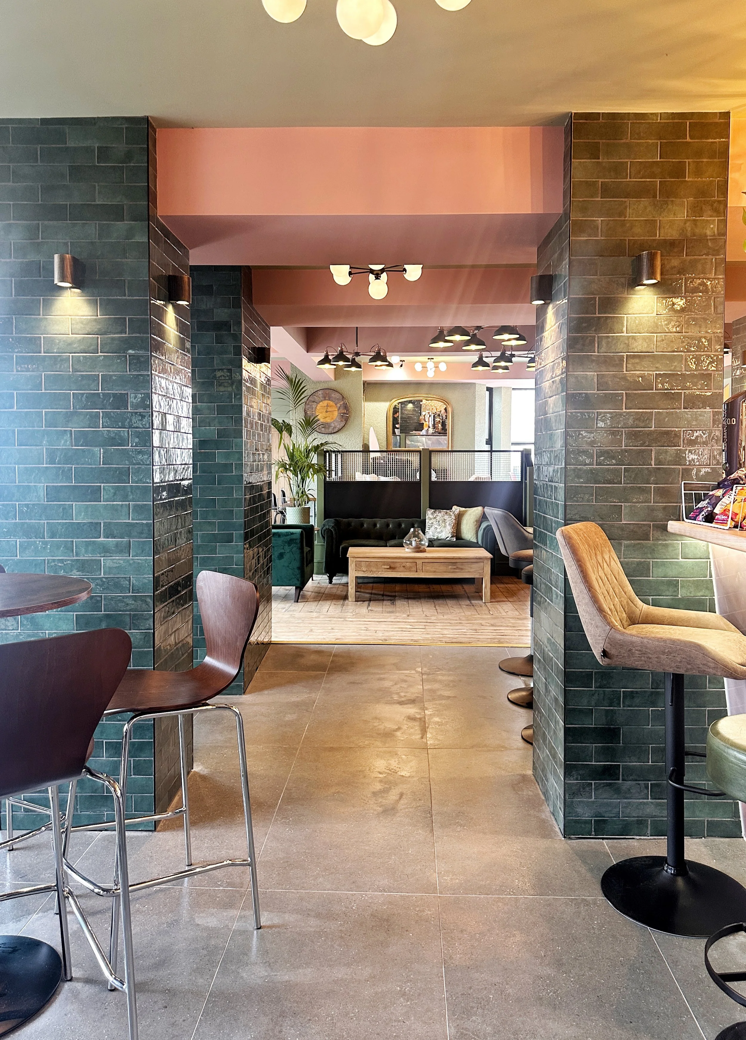 Interior of a modern cafe or lounge with green tiled walls, bar chairs, and a cozy seating area with a sofa and coffee table, decorated with plants and wall art.