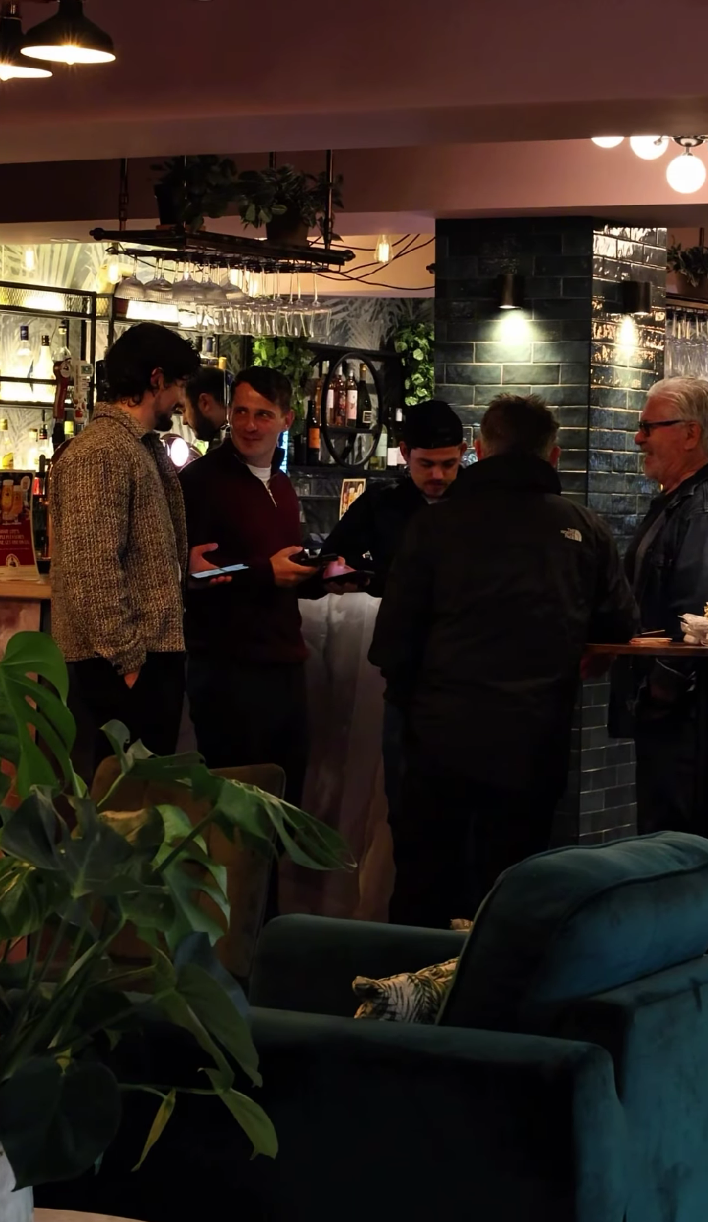 A group of five men gathered at a bar, all holding smartphones, engaged in conversation in a dimly lit social setting with a plant in the foreground and bar elements in the background.