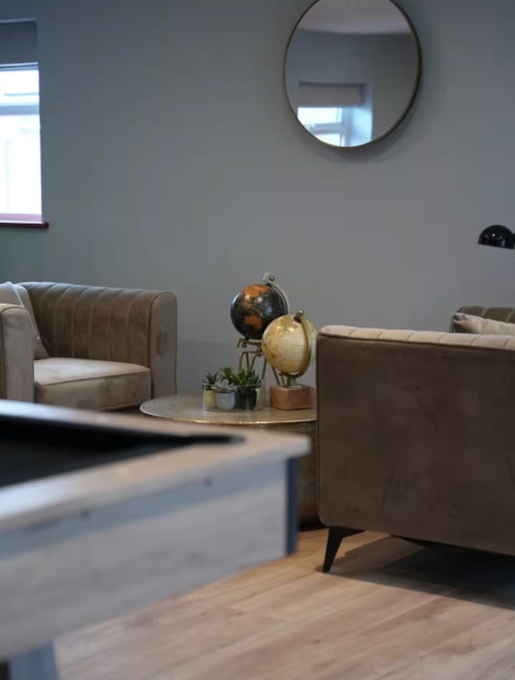 Living room with beige and brown couches, a round coffee table with two globes and a small plant, a large round mirror on the wall, and a window letting in natural light.