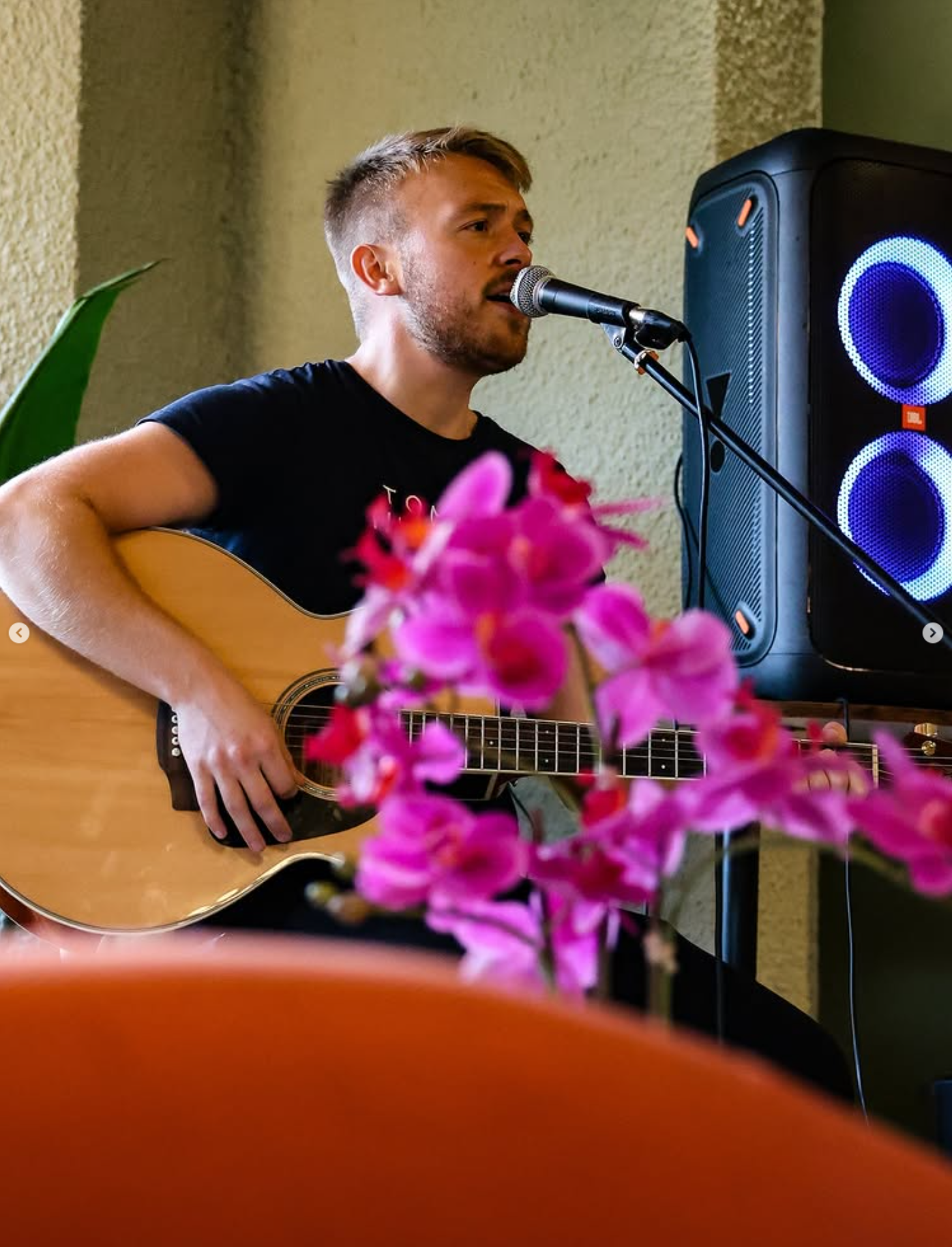 A young man with short hair and a beard playing an acoustic guitar and singing into a microphone, with pink flowers in the foreground and a speaker with blue lights behind him.