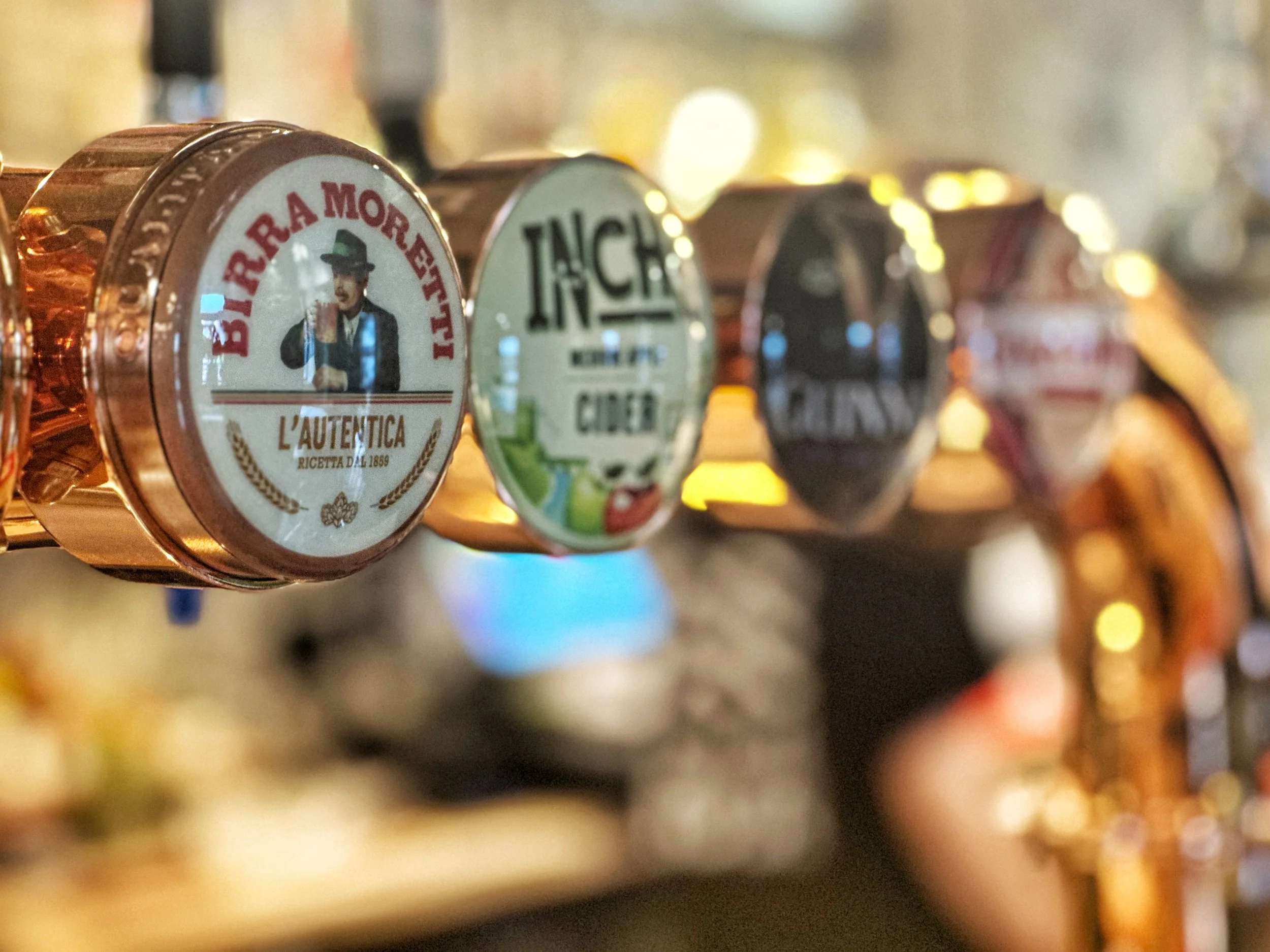 Close-up of beer tap handles, with the front handle featuring a vintage-style label with a man holding a beer, labeled 'Birra Moretti' and 'L'Autentica.' The other handles are blurred in the background.