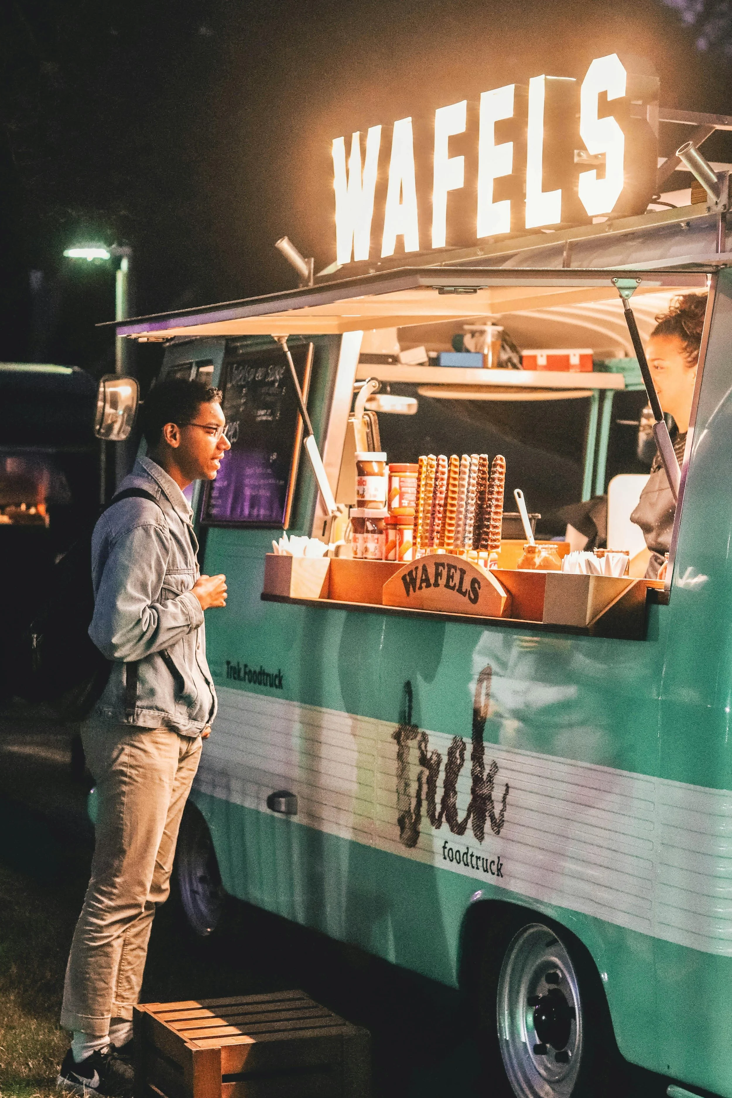 A person in a denim jacket ordering at a teal food truck that sells waffles. The food truck has a bright sign saying 'WAFFLES' and various supplies organized on the counter, with another person inside preparing food.