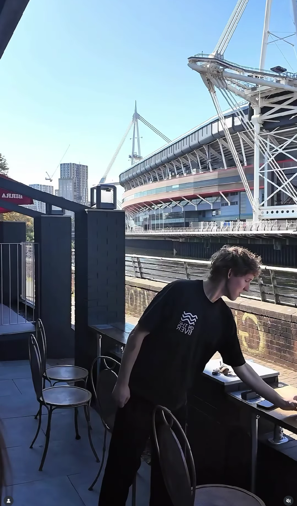 A person standing at a bar table outside, with a large boat or stadium in the background on a sunny day.