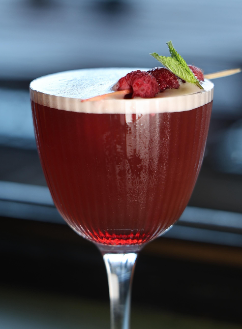 A red cocktail in a stemmed glass garnished with raspberries and a mint leaf.