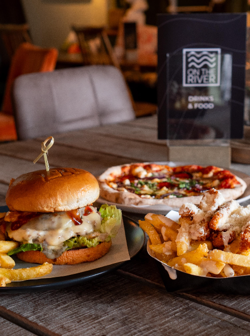 Burger with lettuce, cheese, and sauce, a side of fries, and a pizza with toppings on a wooden table in a dining setting.