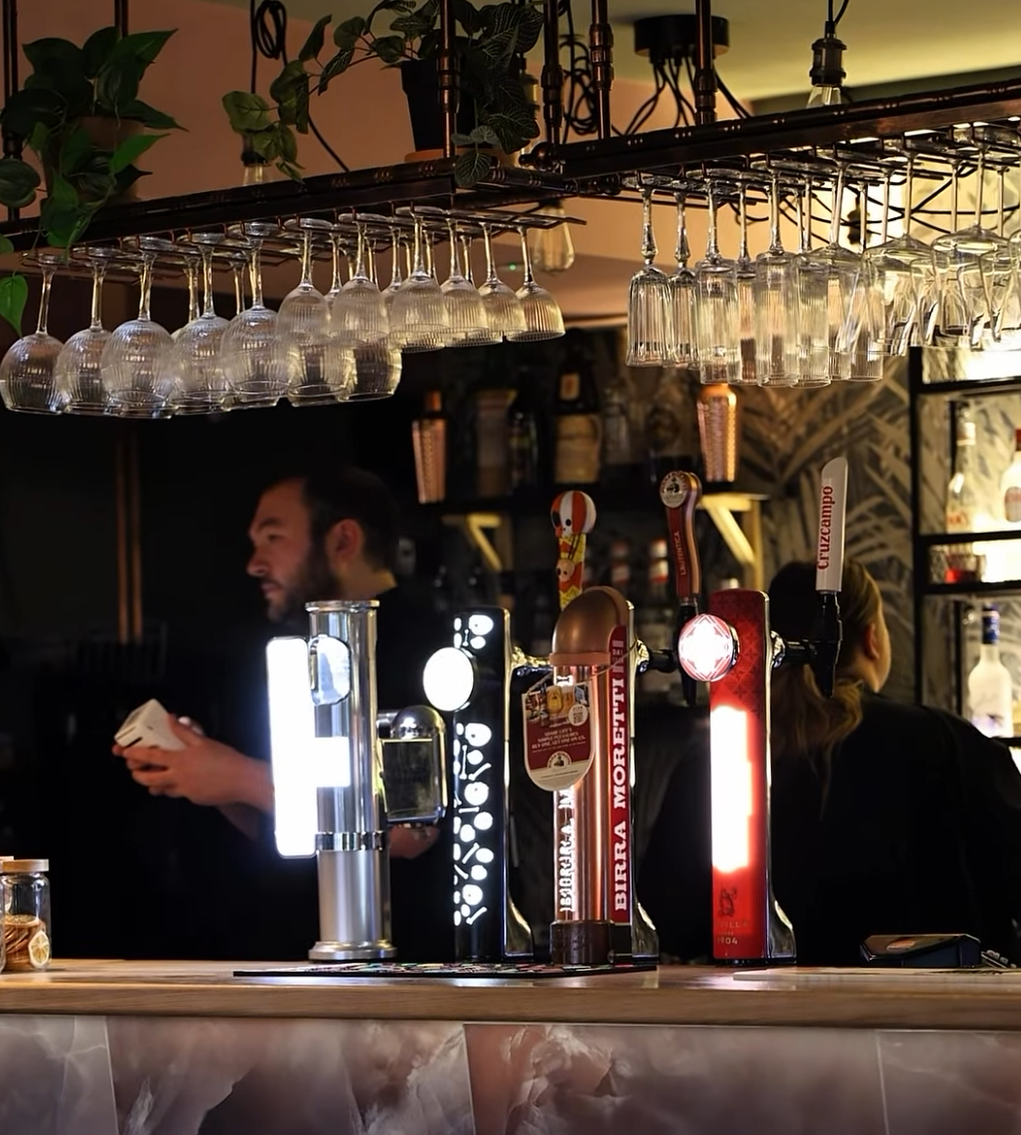 A bar with beer taps, hanging glasses, and two bartenders. The background features plants, shelves, and decorative wallpaper.